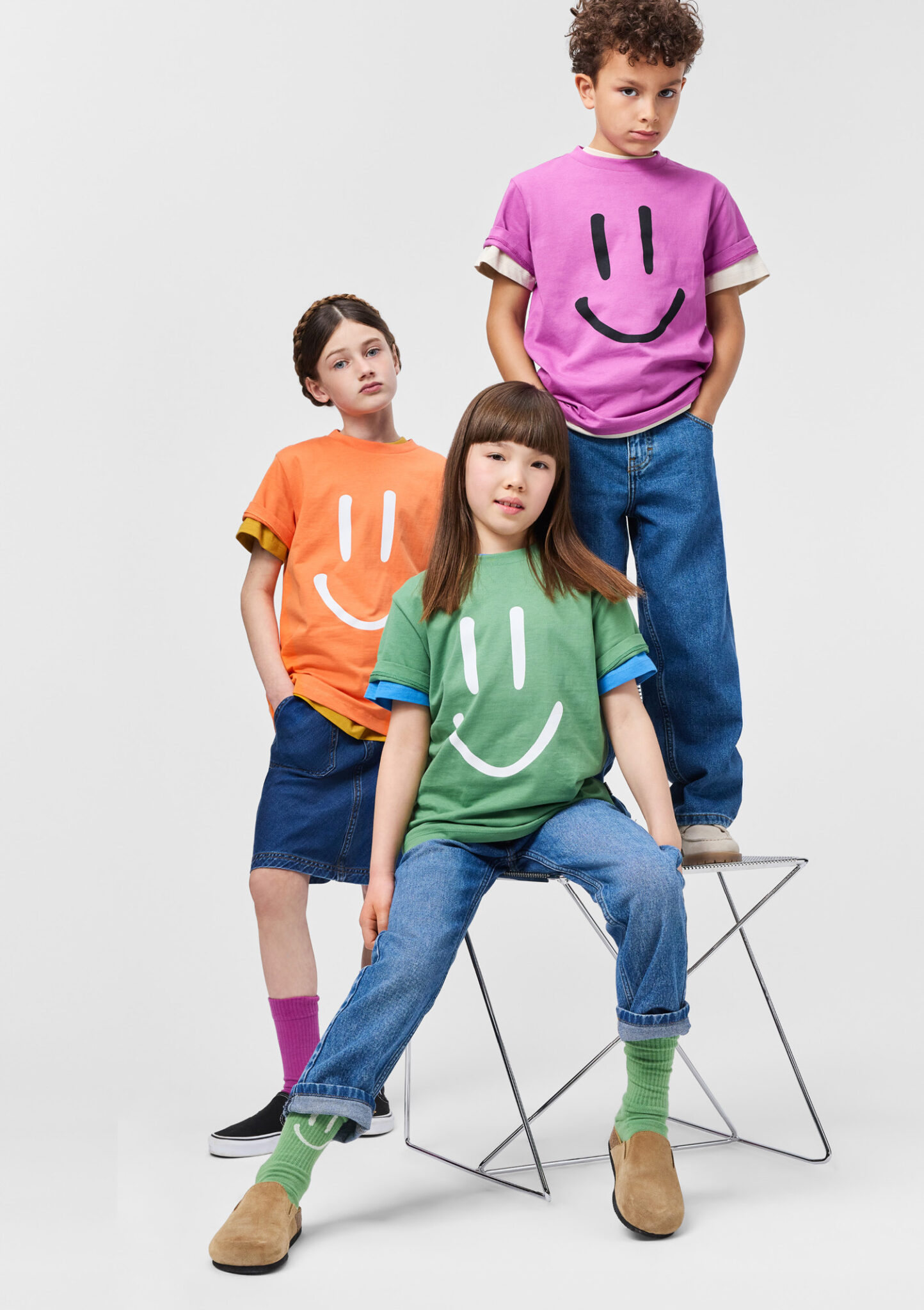 Three kids wearing colorful smiley face T-shirts from Molo SS26, studio group portrait with bold graphic kidswear featured in Scimparello Magazine.