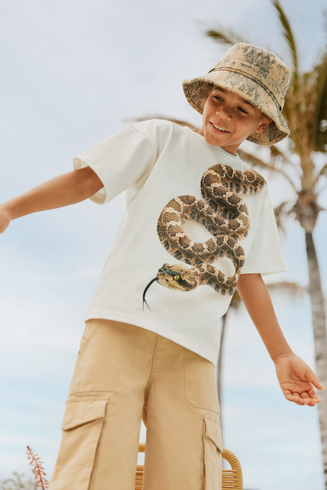 Boy wearing snake print T-shirt and beige cargo shorts from Molo SS26, outdoor summer look featured in Scimparello Magazine.