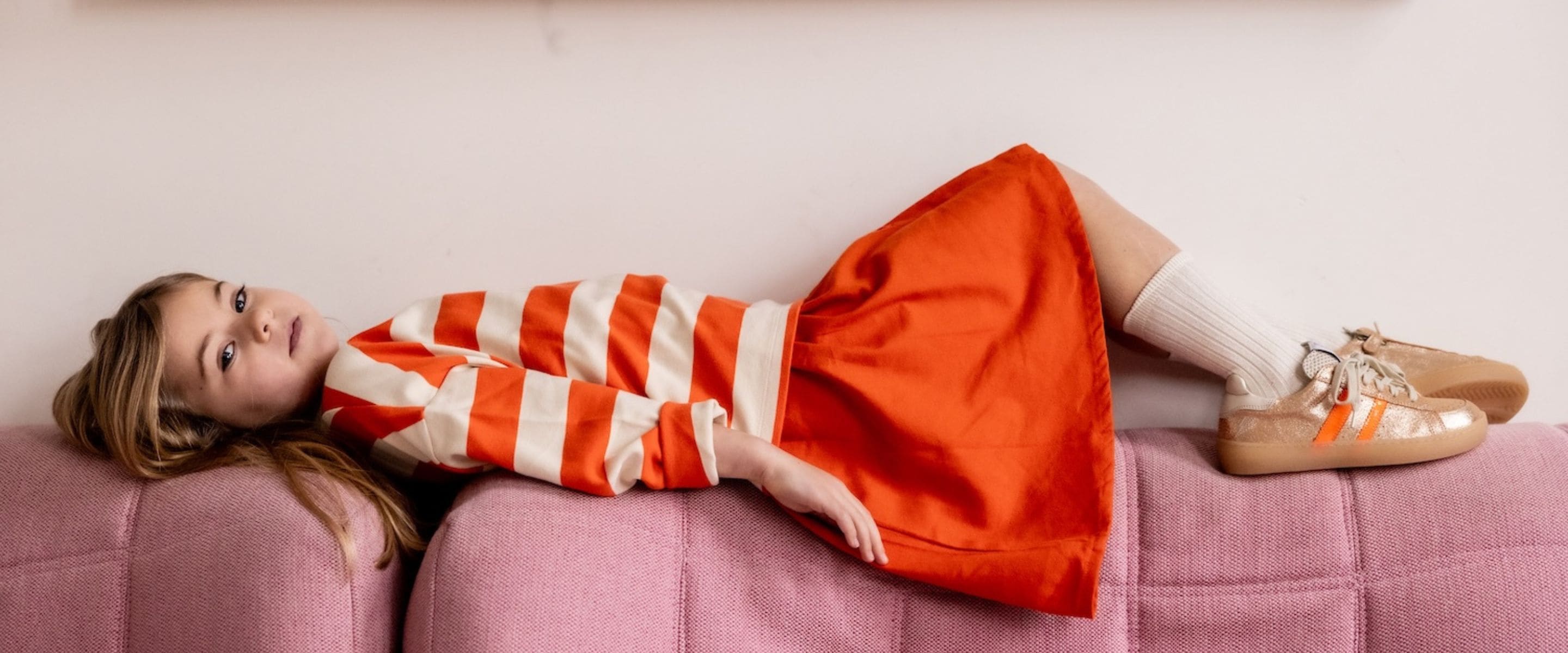 Girl lying sideways on a pink upholstered bench wearing an orange and cream striped long-sleeve top, bright orange skirt, white ribbed socks and gold sneakers with orange stripes from Ocra SS26, featured in Scimparello Magazine.