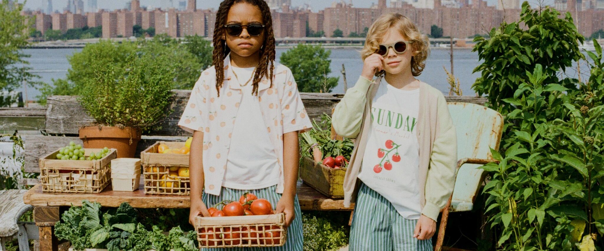 The Sunday Collective kidswear image featuring two children wearing fruit-print shirts, striped shorts and sunglasses, standing by wooden crates filled with fresh tomatoes and citrus on a rooftop garden overlooking the city – Scimparello Magazine.
