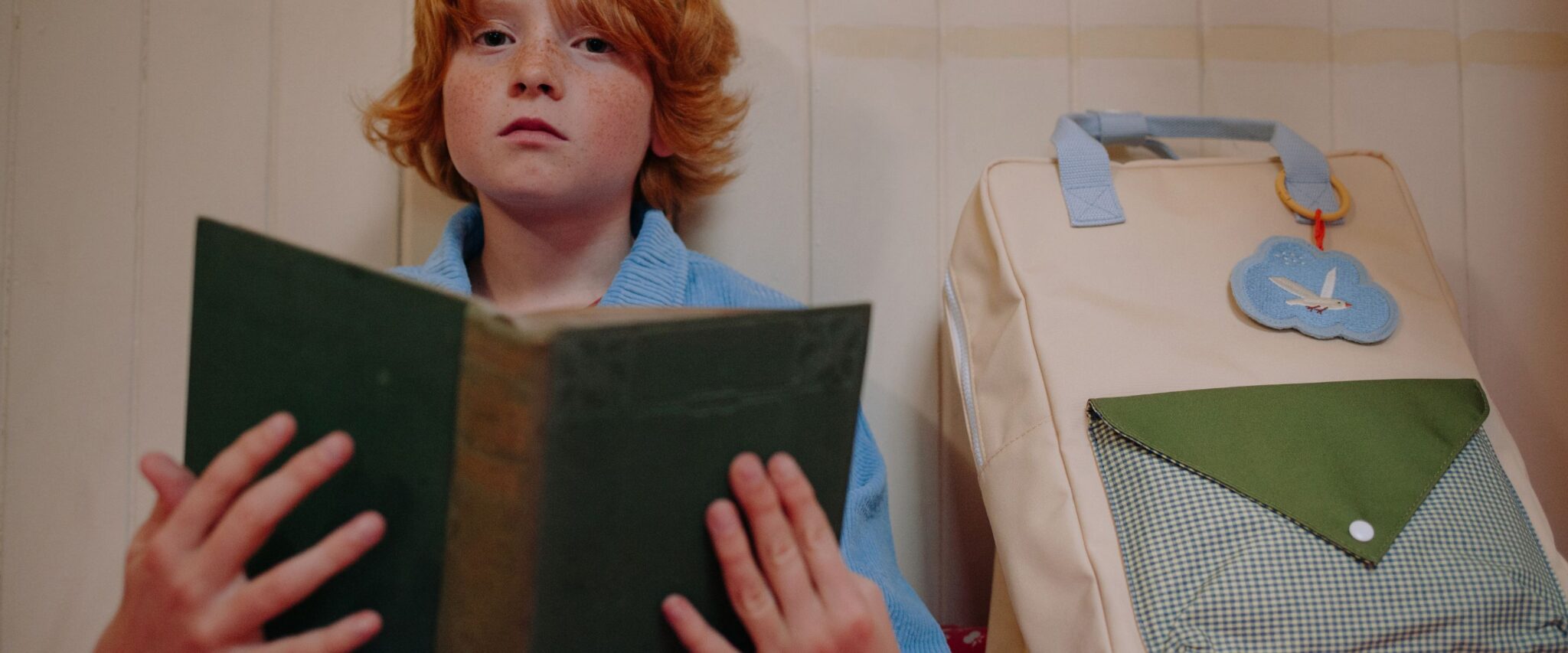 Sticky Lemon SS26 kids collection school backpack in cream with green flap and gingham pocket, blue cloud patch detail, styled beside a red-haired child reading a green book indoors – Scimparello Magazine.