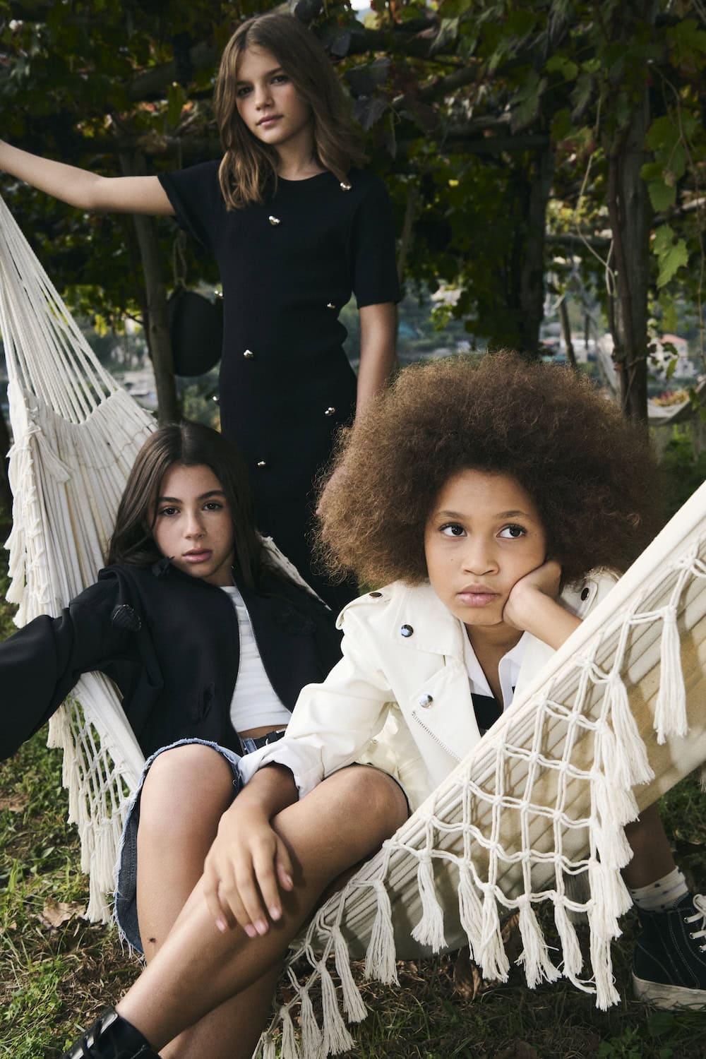 Close-up of three girls posing in a hammock surrounded by greenery, wearing black minimalist dresses and a white jacket with sporty details, To Be Too SS26 summer collection featured in Scimparello Magazine.