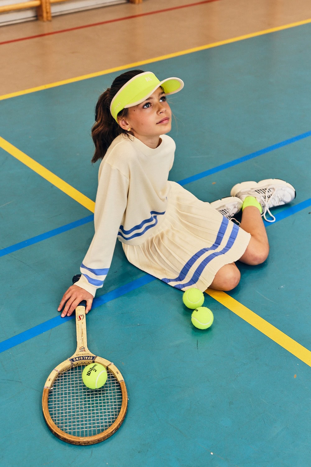 Girl wearing AO76 Spring Summer 26 collection sitting on indoor gym floor with vintage tennis racket and balls, pleated knit dress with blue stripes and neon visor – Scimparello Magazine Collections SS26.