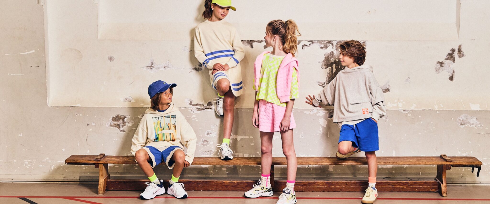 Kids wearing AO76 Spring Summer 26 collection in sporty pastel outfits with hoodies, shorts and sneakers, standing on a wooden bench against a textured wall – Scimparello Magazine Collections SS26.