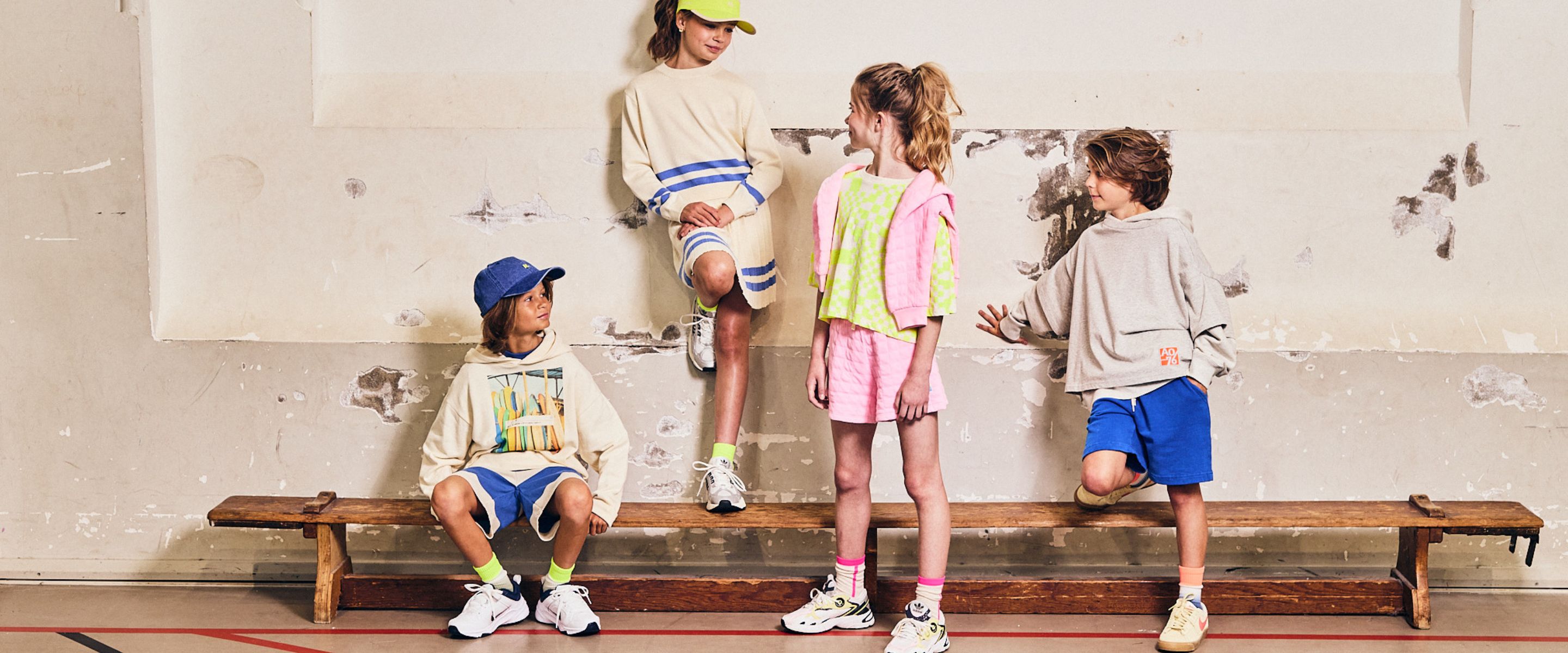 Kids wearing AO76 Spring Summer 26 collection in sporty pastel outfits with hoodies, shorts and sneakers, standing on a wooden bench against a textured wall – Scimparello Magazine Collections SS26.