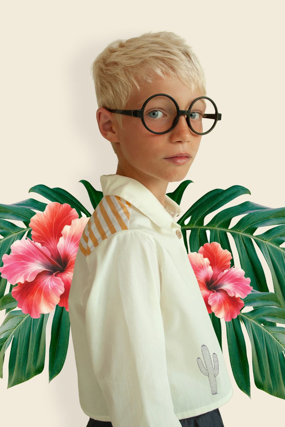Blond boy wearing round black glasses and a cream shirt with striped shoulder detail and cactus embroidery, posed in front of tropical green leaves and pink hibiscus flowers – Bianca & Noè SS26 Collection featured on Scimparello Magazine.