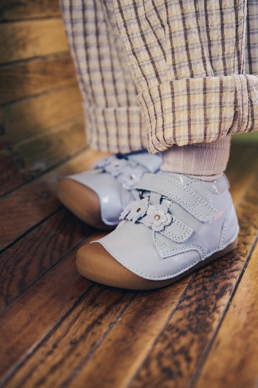 Bundgaard SS26 light blue leather toddler shoes with double velcro straps and floral detail, worn with checked trousers on a wooden floor in a vintage travel setting, featured in Scimparello Magazine.