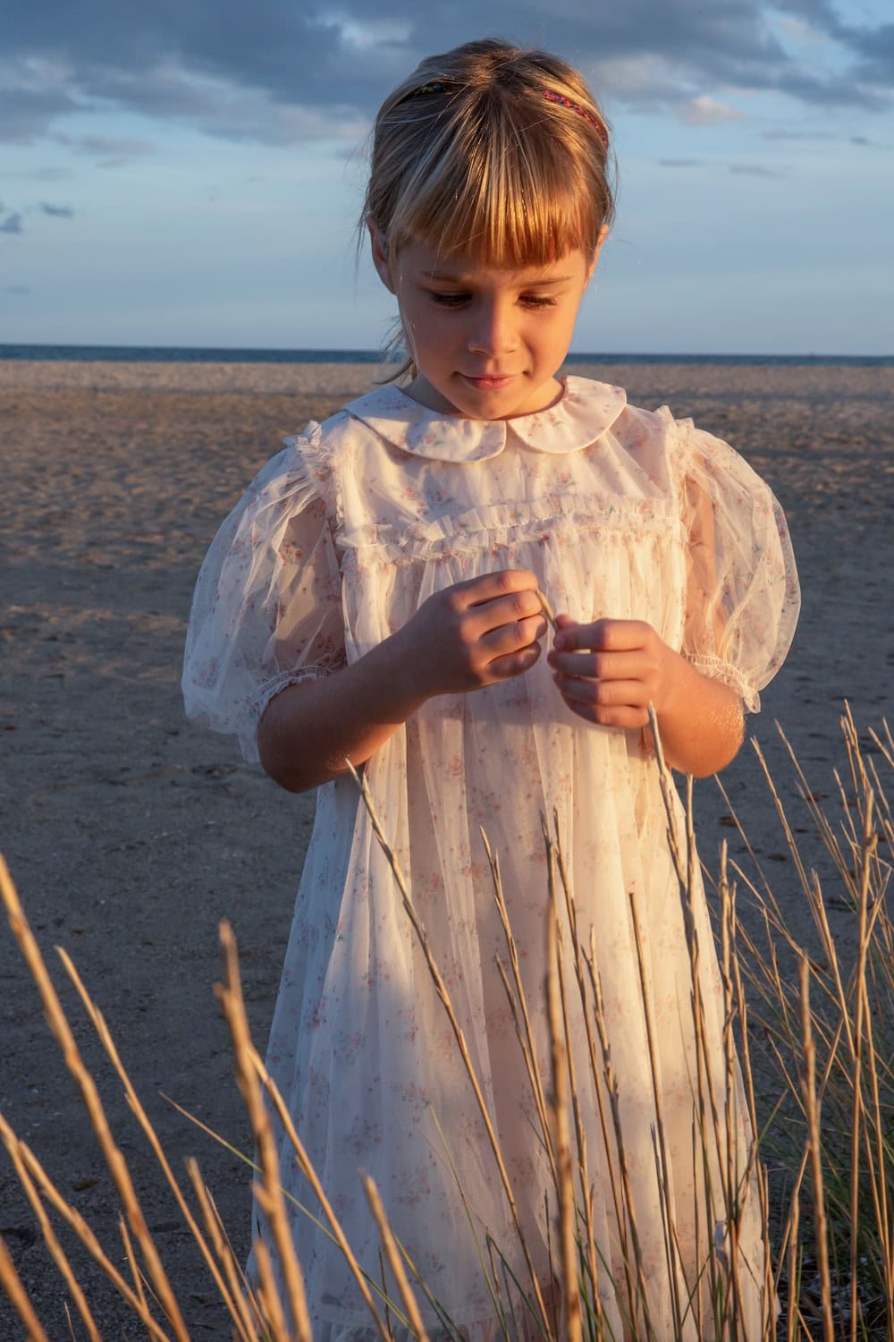 Girl wearing a delicate floral tulle dress with puff sleeves and a Peter Pan collar, standing on a sandy beach at sunset holding dry grass stems, photographed in golden light for C’era Una Volta SS26 featured in Scimparello Magazine.