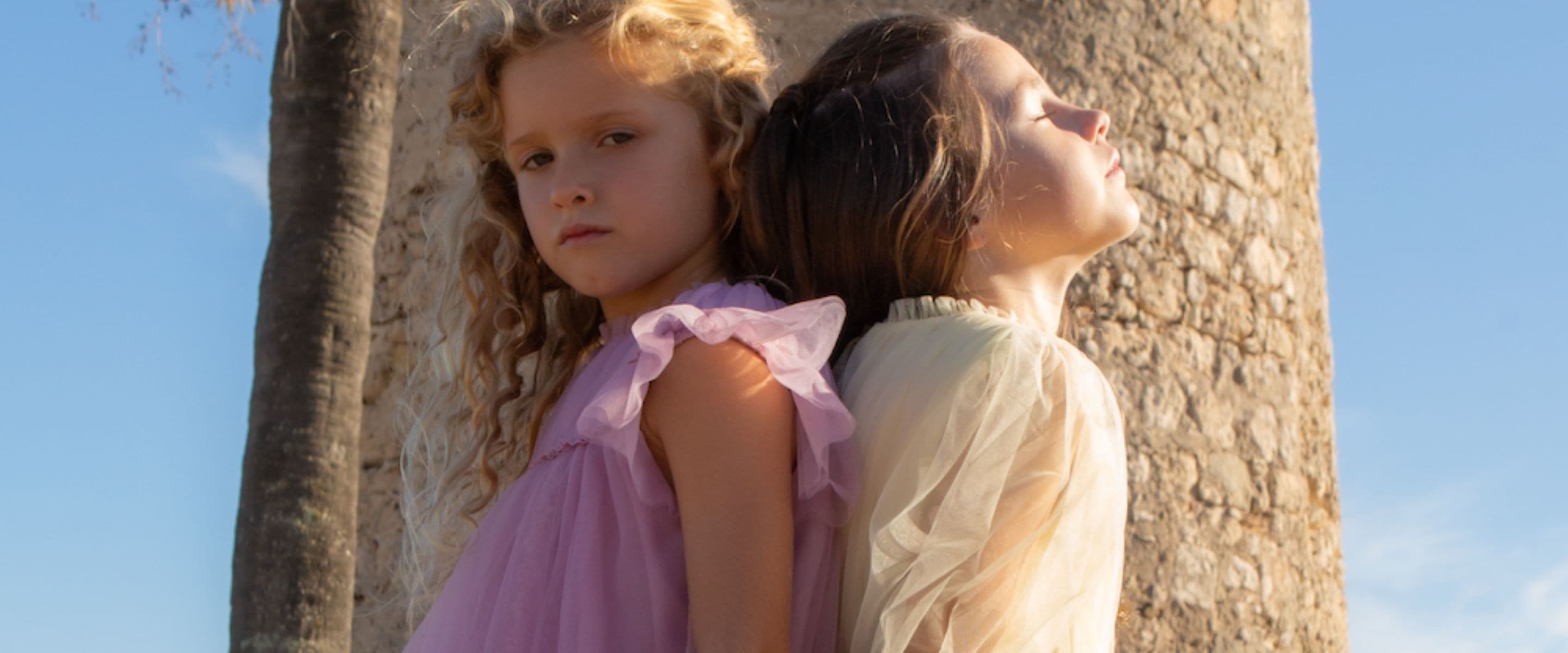Two girls standing back to back against a sunlit stone wall, wearing soft pastel dresses in lilac and cream with delicate ruffle details, photographed in warm golden light for C’era Una Volta SS26 – Scimparello Magazine.
