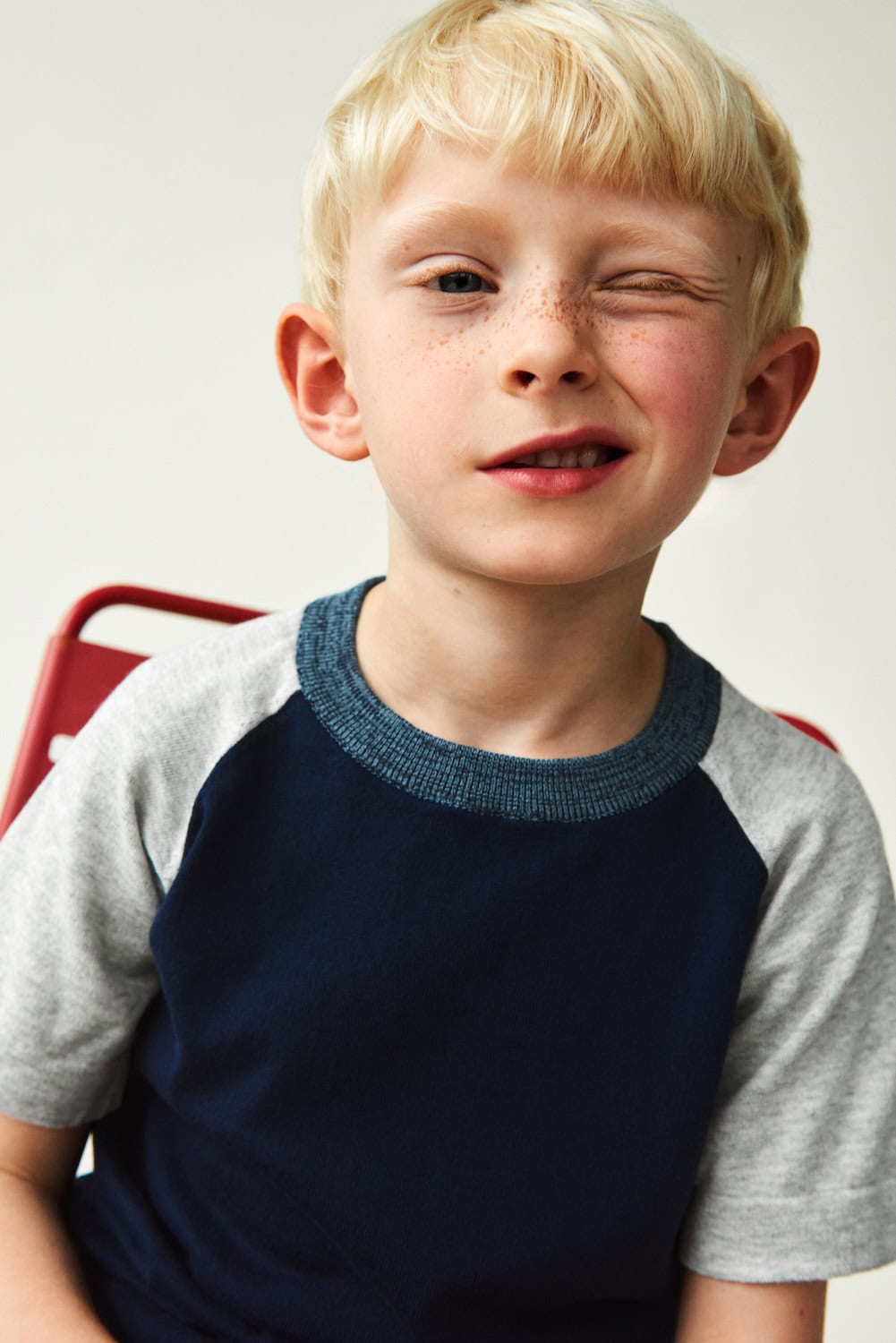 FUB Kids SS26 – close-up portrait of blond boy winking, wearing a navy and grey raglan knit top, playful studio moment, featured on Scimparello Magazine.