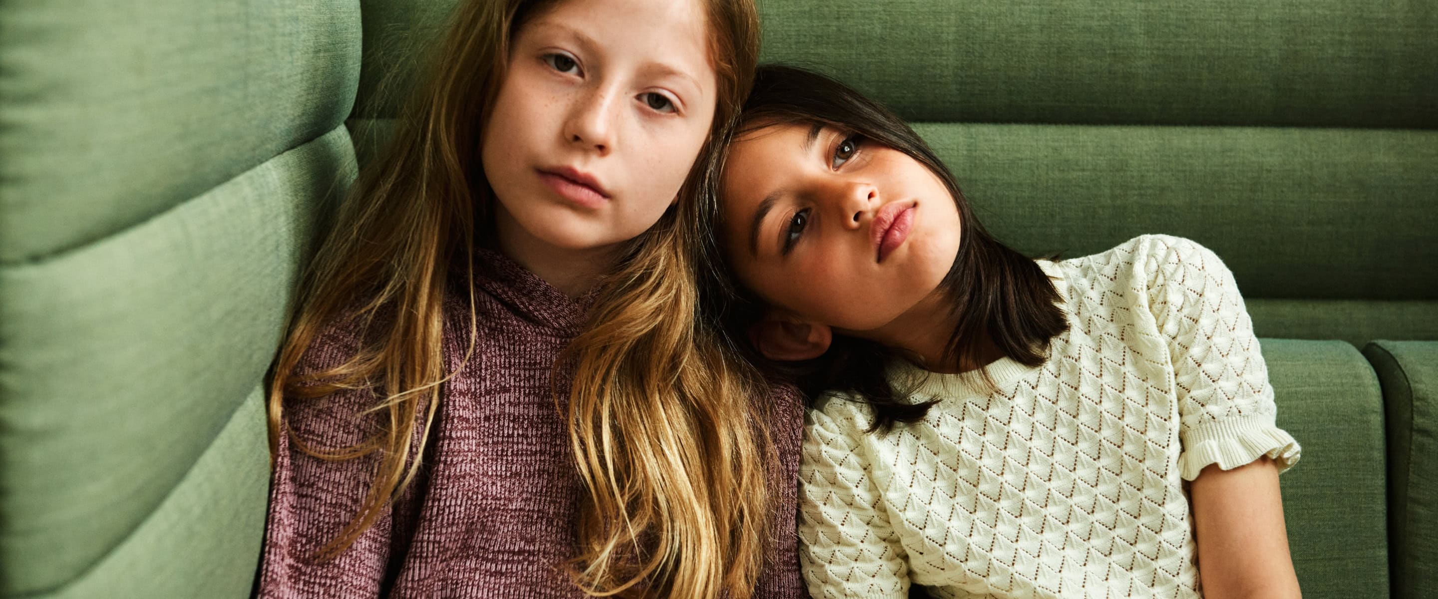 FUB Kids SS26 collection – two girls in textured knit tops in soft plum and cream tones leaning against a green upholstered sofa, natural light portrait, minimalist Nordic aesthetic, featured on Scimparello Magazine.