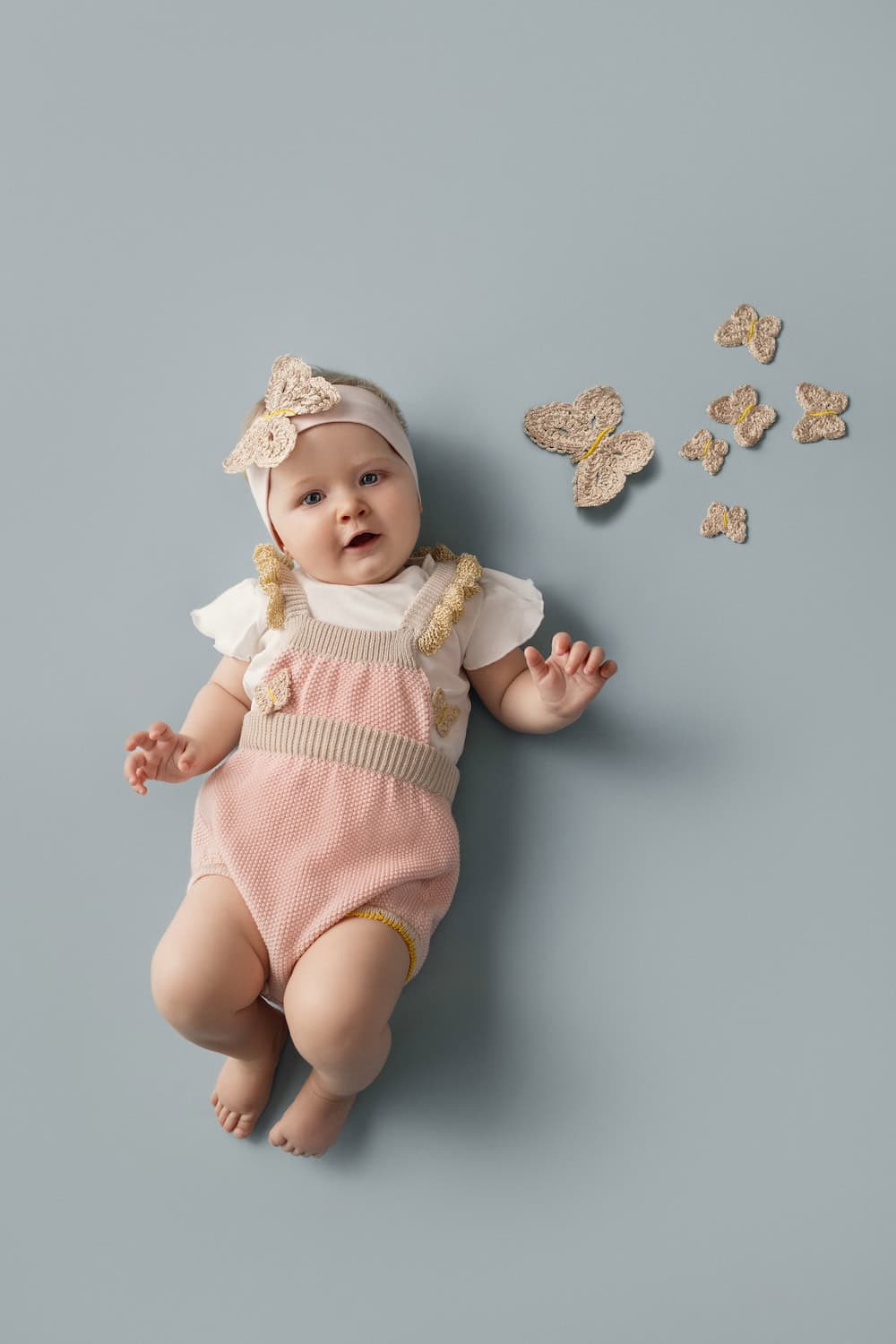 Baby girl lying on a soft blue background wearing a pink knit romper with beige trims and crochet butterfly details from the Gensami SS26 baby collection – Scimparello Magazine.
