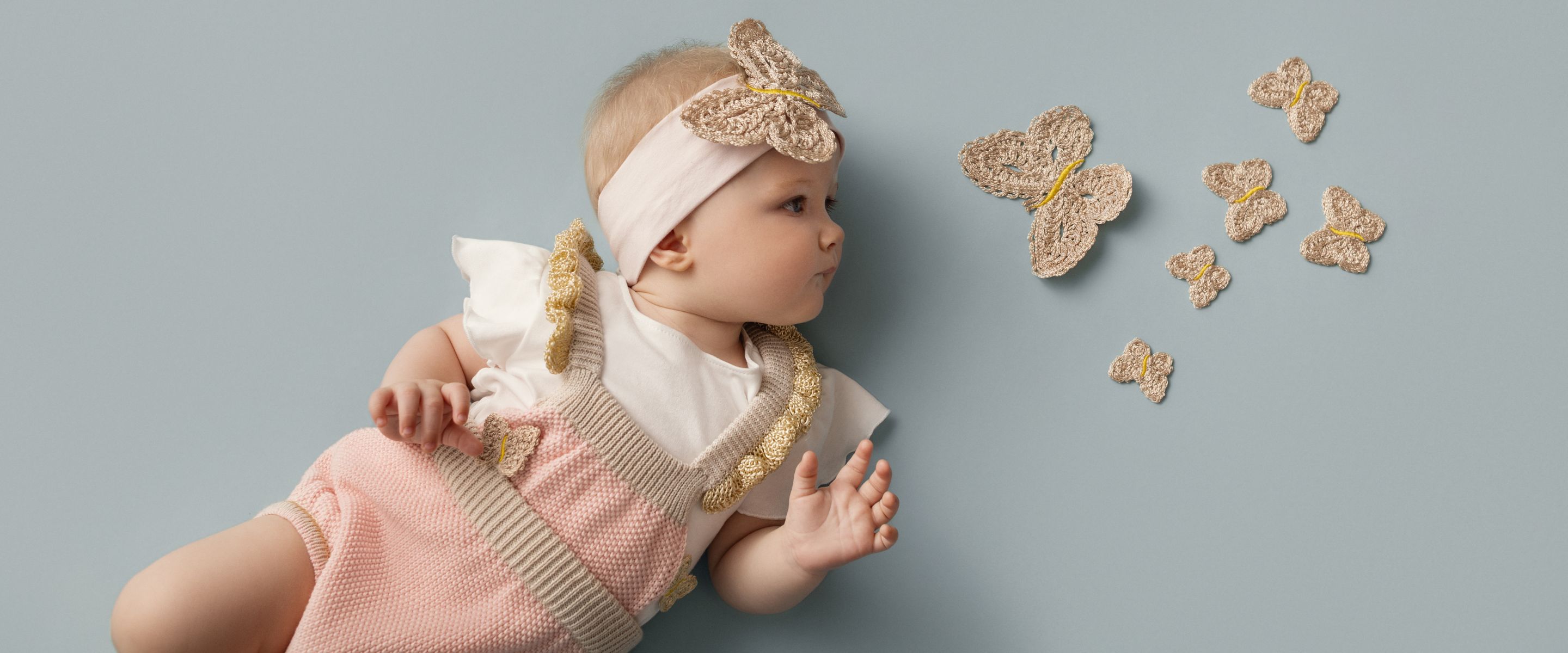 Baby girl wearing a soft pink knitted romper with beige trim and crochet butterfly details, styled with a matching headband, photographed against a light blue background for Gensami SS26 – Scimparello Magazine.