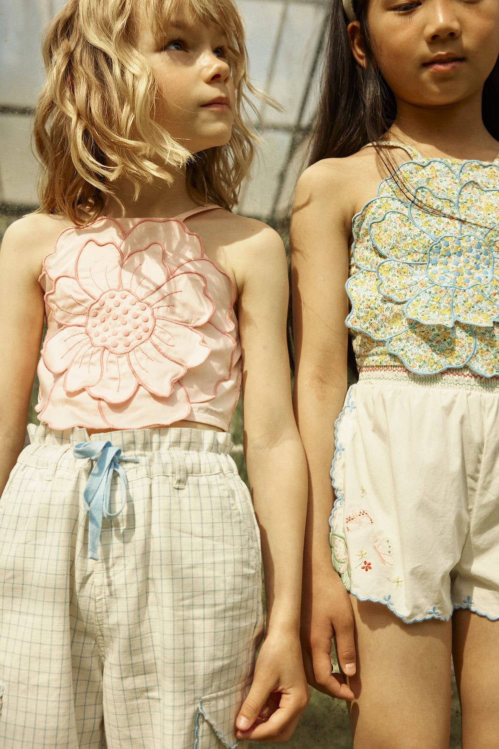 Close-up of two girls wearing pastel flower-appliqué tops paired with embroidered shorts and checked trousers, Gingersnaps SS26 – Scimparello Magazine