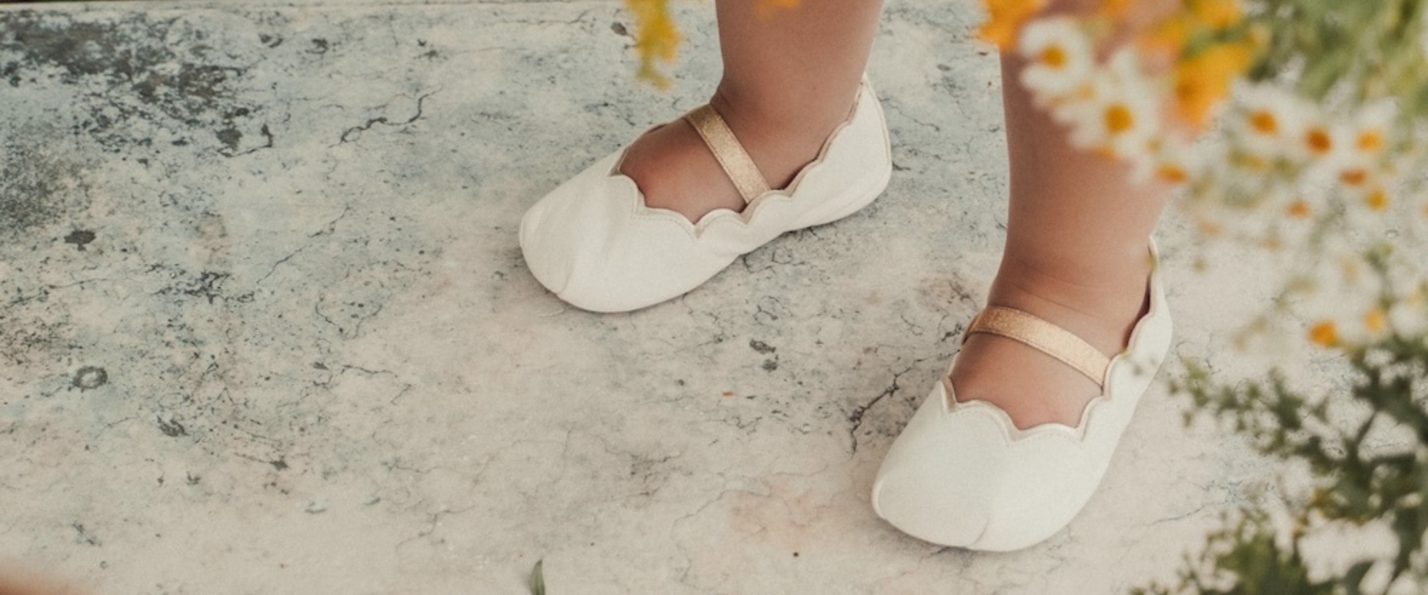 Pèpè Children Shoes SS26 white leather Mary Jane flats with scalloped edges and gold strap detail on a child standing on natural stone, soft floral background, featured on Scimparello Magazine.
