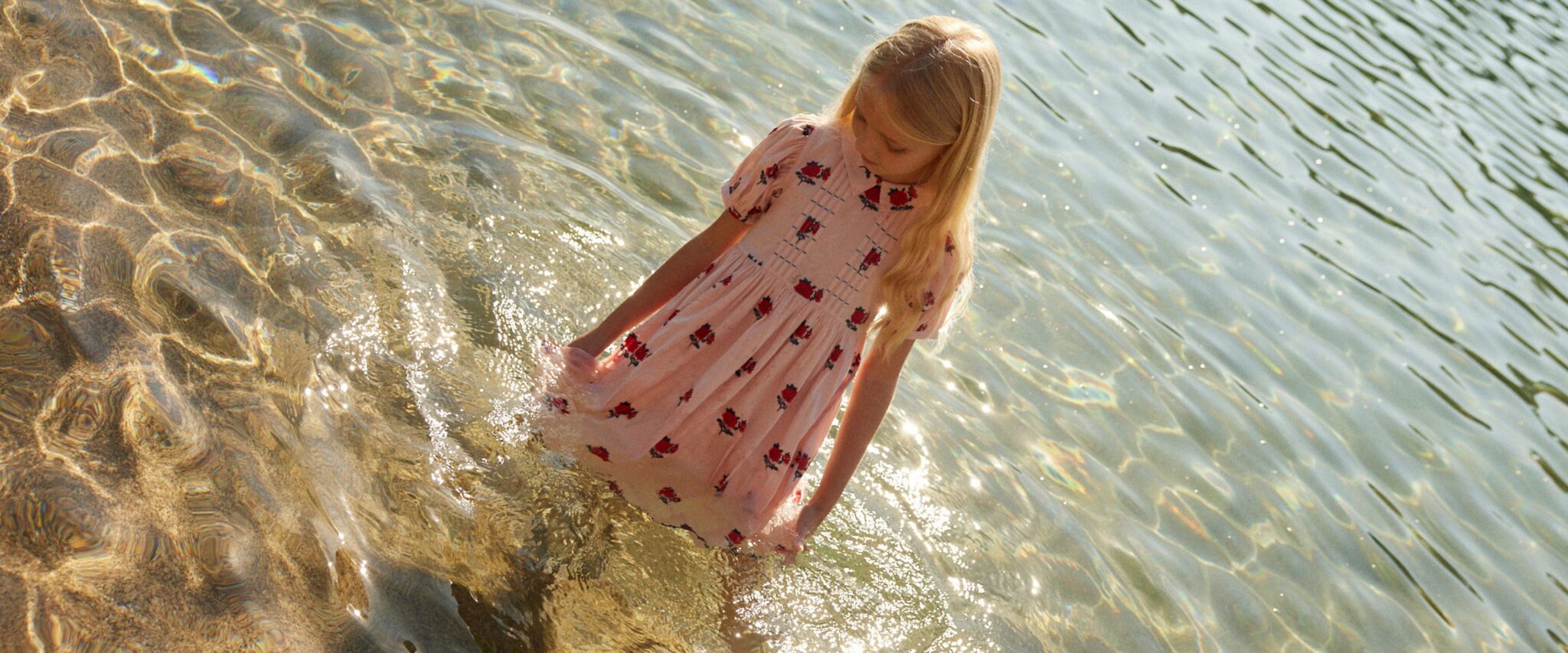Bebe Organic SS26 girl wearing a soft pink floral dress standing in shallow sunlit water, golden reflections and ripples around her feet, romantic seaside mood, featured on Scimparello Magazine.