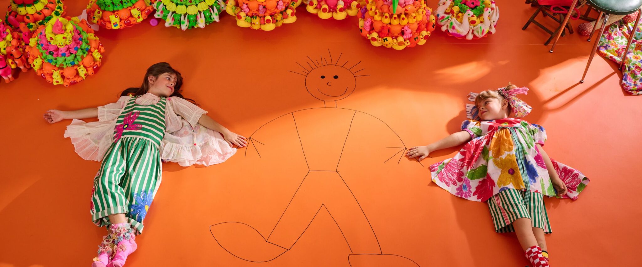 Two girls wearing colorful striped and floral dresses from Péro SS26 lying on bright orange floor beside hand-drawn figure, playful kids fashion scene featured in Scimparello Magazine.