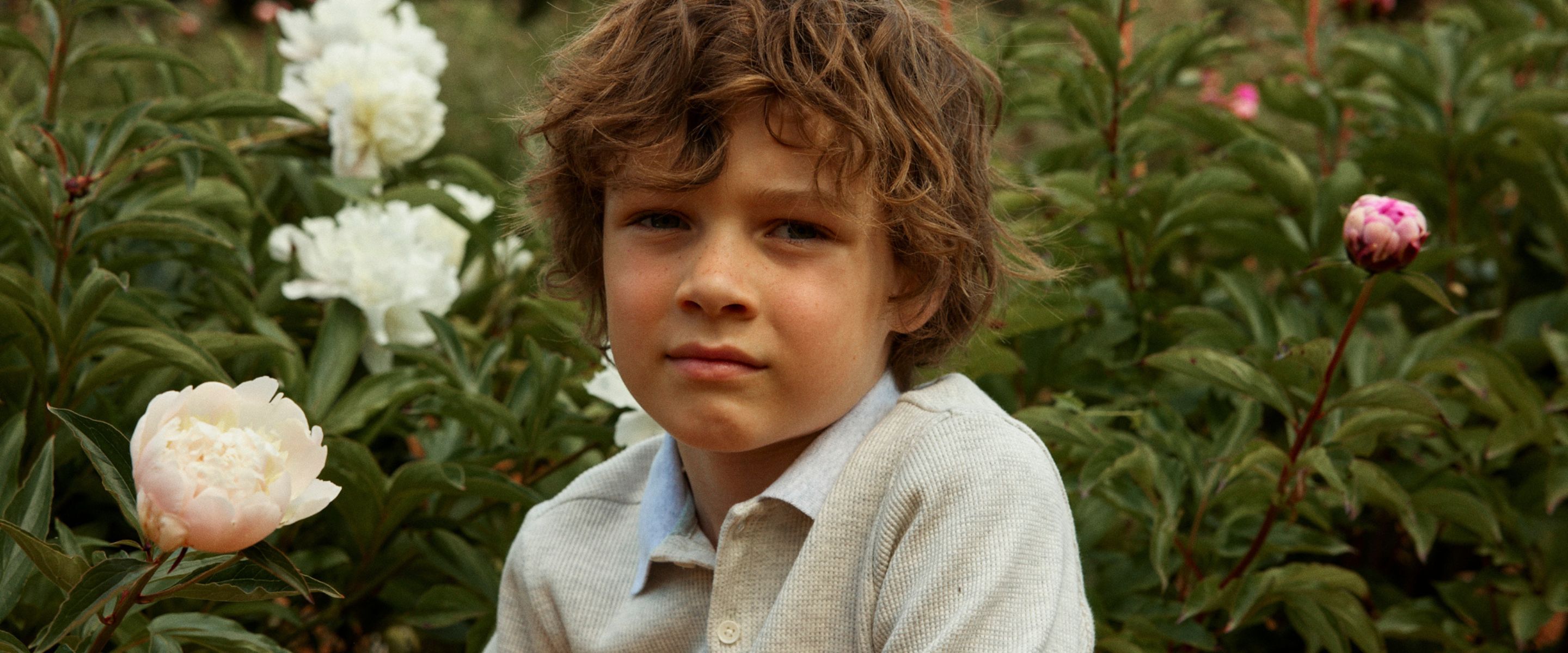 Boy with curly brown hair wearing a beige knit polo shirt surrounded by blooming peonies in a garden, Gingersnaps SS26 collection – Scimparello Magazine