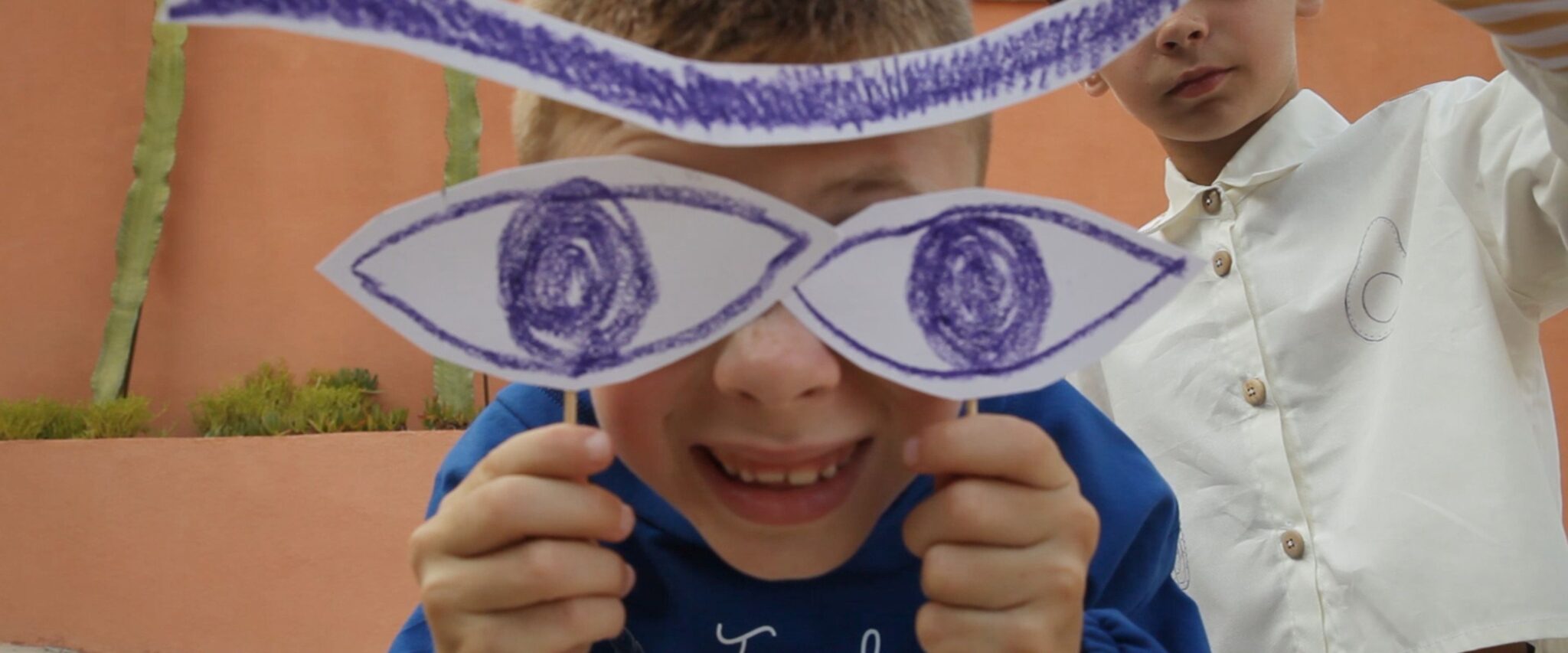 Close-up of a boy holding hand-drawn paper eyes in front of his face, second child in white shirt behind him against a terracotta wall – Bianca & Noè SS26 Collection on Scimparello Magazine.