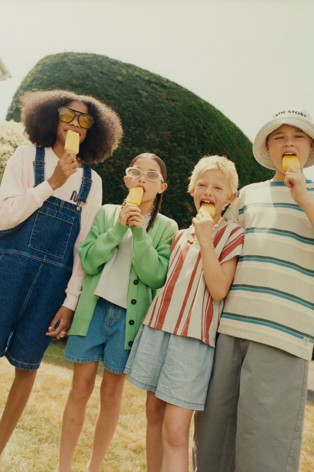 Four children eating yellow ice pops in a sunny garden, dressed in denim and striped summer looks, MAIN STORY SS26 on Scimparello Magazine.