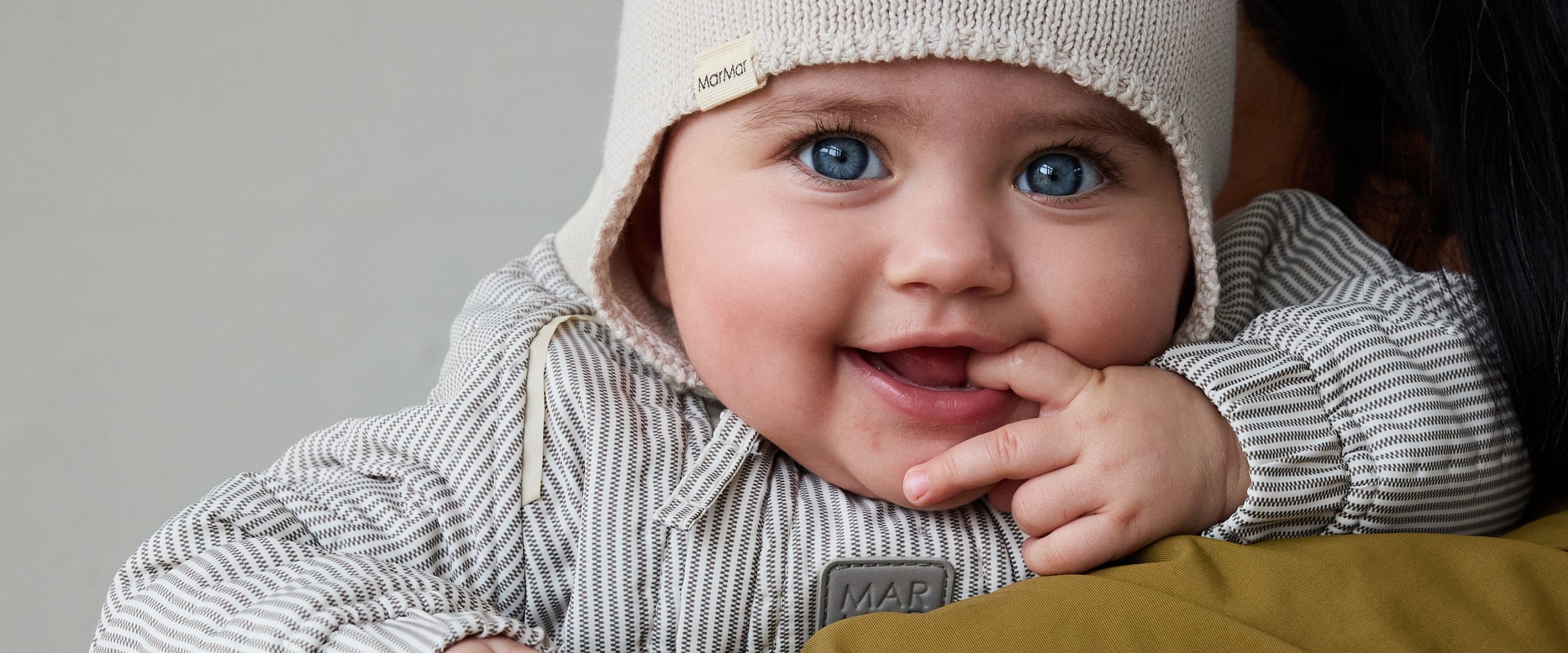 MarMar Copenhagen SS26 baby collection featured in Scimparello Magazine – close-up portrait of a smiling baby with bright blue eyes wearing a cream knitted bonnet and a beige striped cotton outfit with hood, soft neutral tones and natural light background.