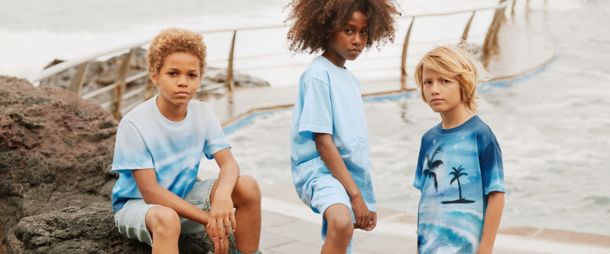 Three boys standing on coastal rocks wearing blue tie-dye and ocean-inspired graphic T-shirts with matching shorts, photographed by the sea for Molo SS26 featured in Scimparello Magazine.
