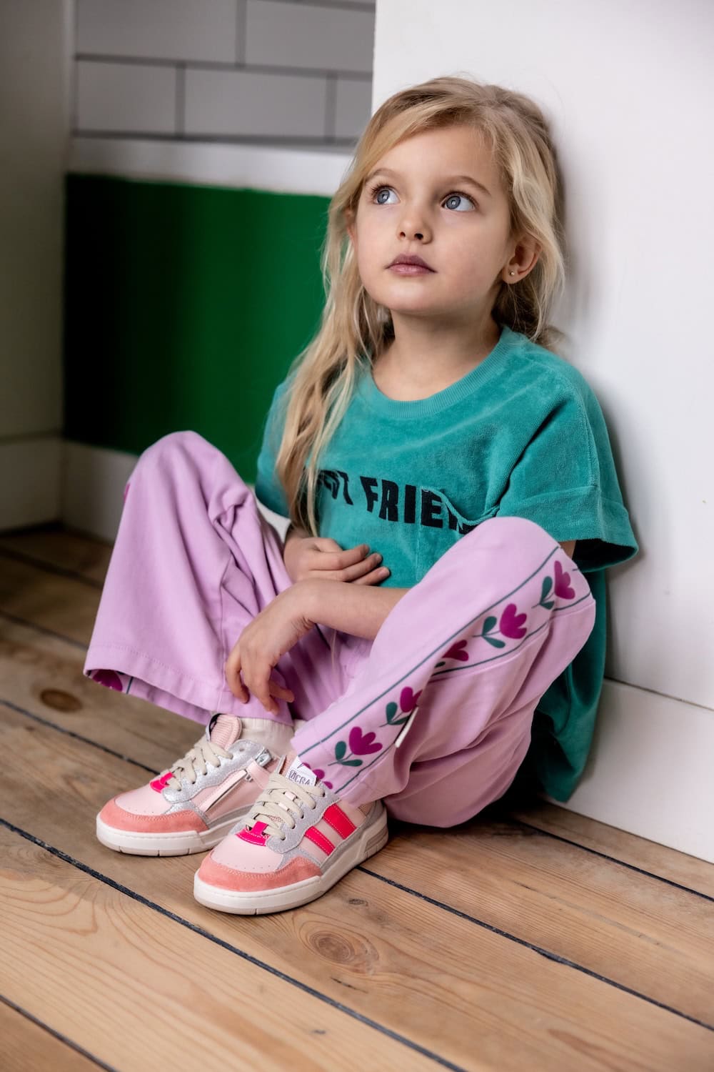 Girl sitting on wooden floor wearing pink and cream sneakers with bright pink stripes from Ocra SS26, paired with lilac pants and green T-shirt, featured in Scimparello Magazine.