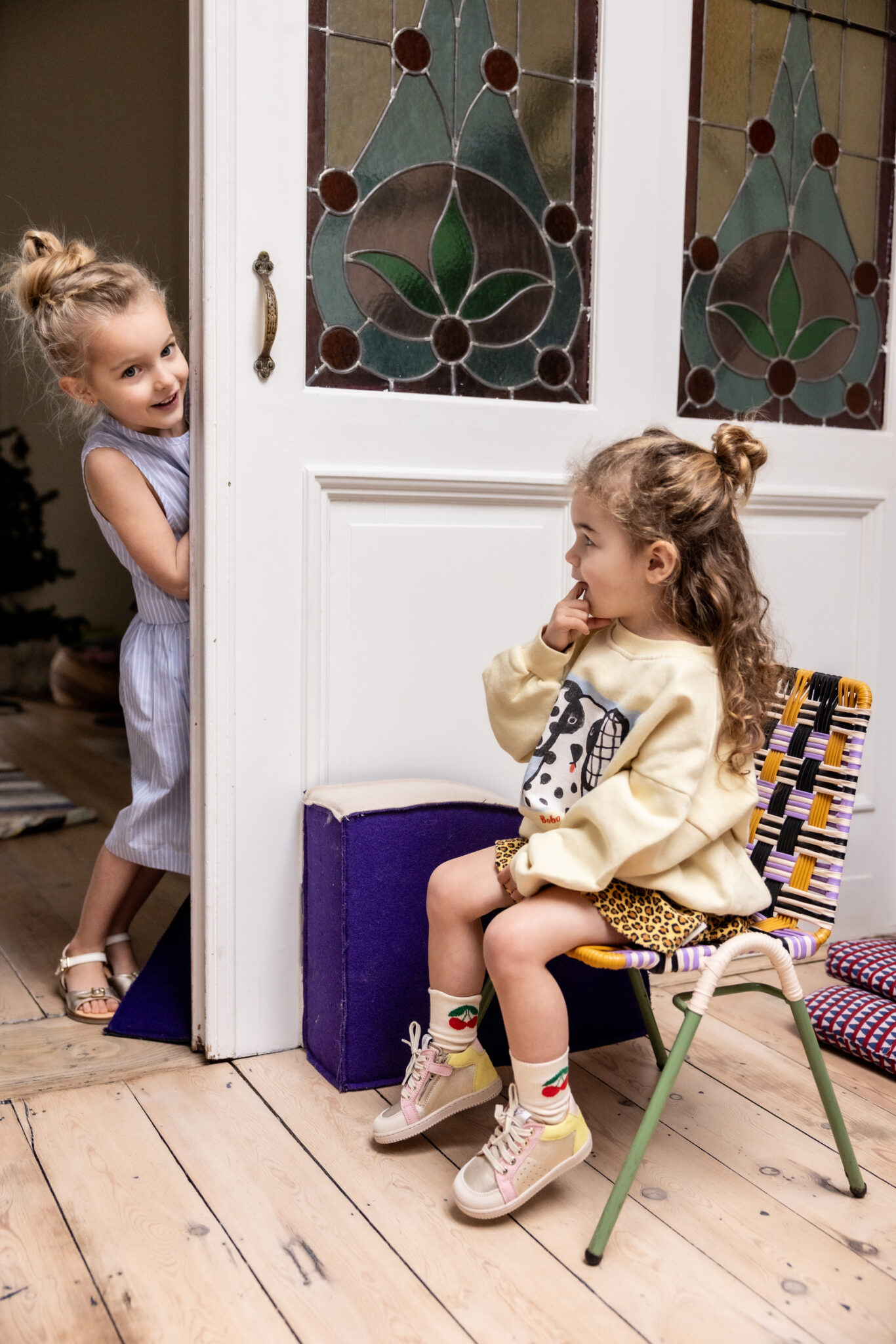 Two girls indoors, one peeking through a door, wearing Ocra SS26 children’s shoes including gold sandals and pink sneakers, featured in Scimparello Magazine.