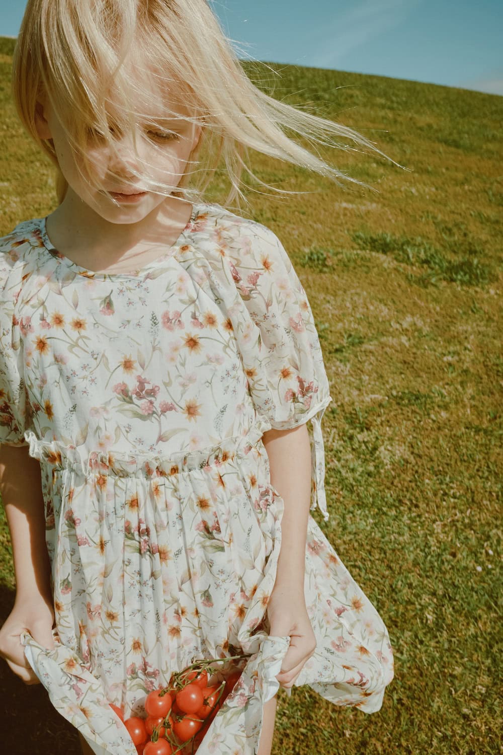 Blonde girl wearing a soft cream floral dress holding cherry tomatoes in the fabric of her skirt, standing on a sunlit grassy hill, Paade Mode SS26 featured in Scimparello Magazine.