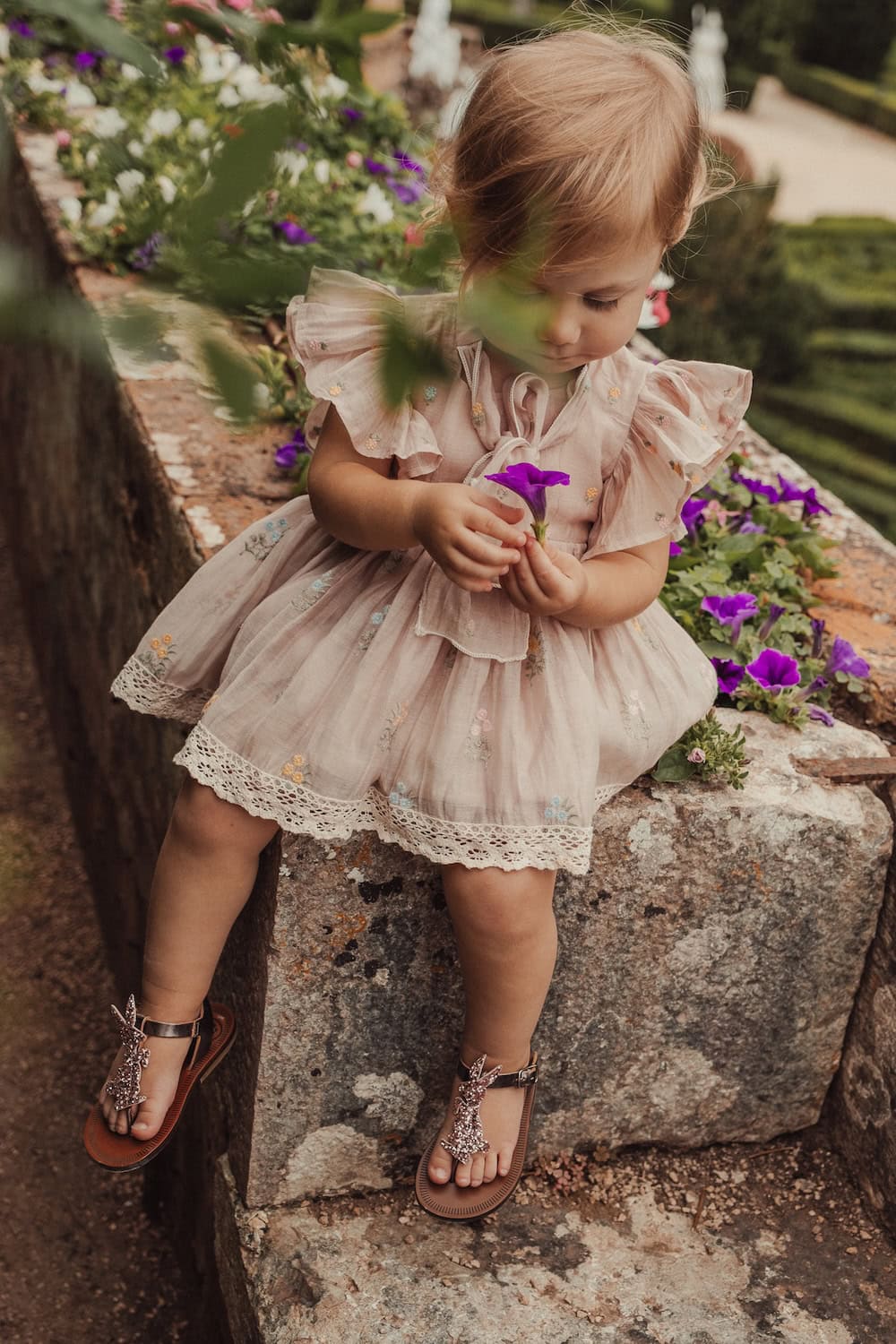 Toddler wearing Pèpè Children Shoes SS26 metallic bronze sandals with glitter dinosaur appliqué, seated on stone in a garden setting, featured on Scimparello Magazine.