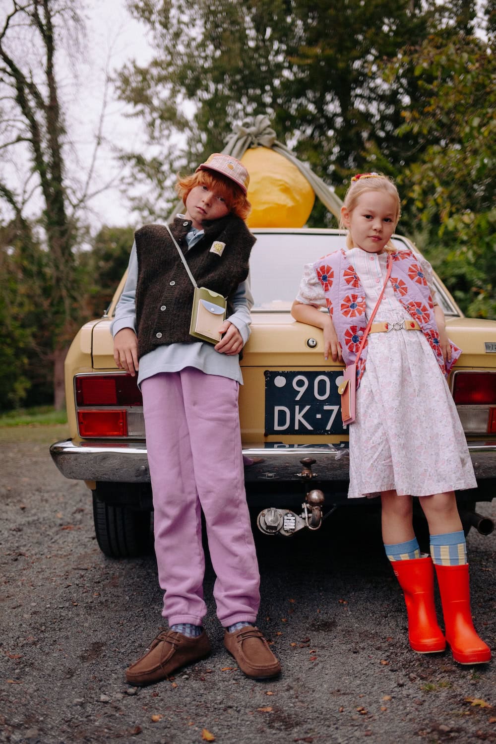 Sticky Lemon SS26 kids collection crossbody phone pouch in pastel pink and yellow worn by child leaning against vintage car – Scimparello Magazine.