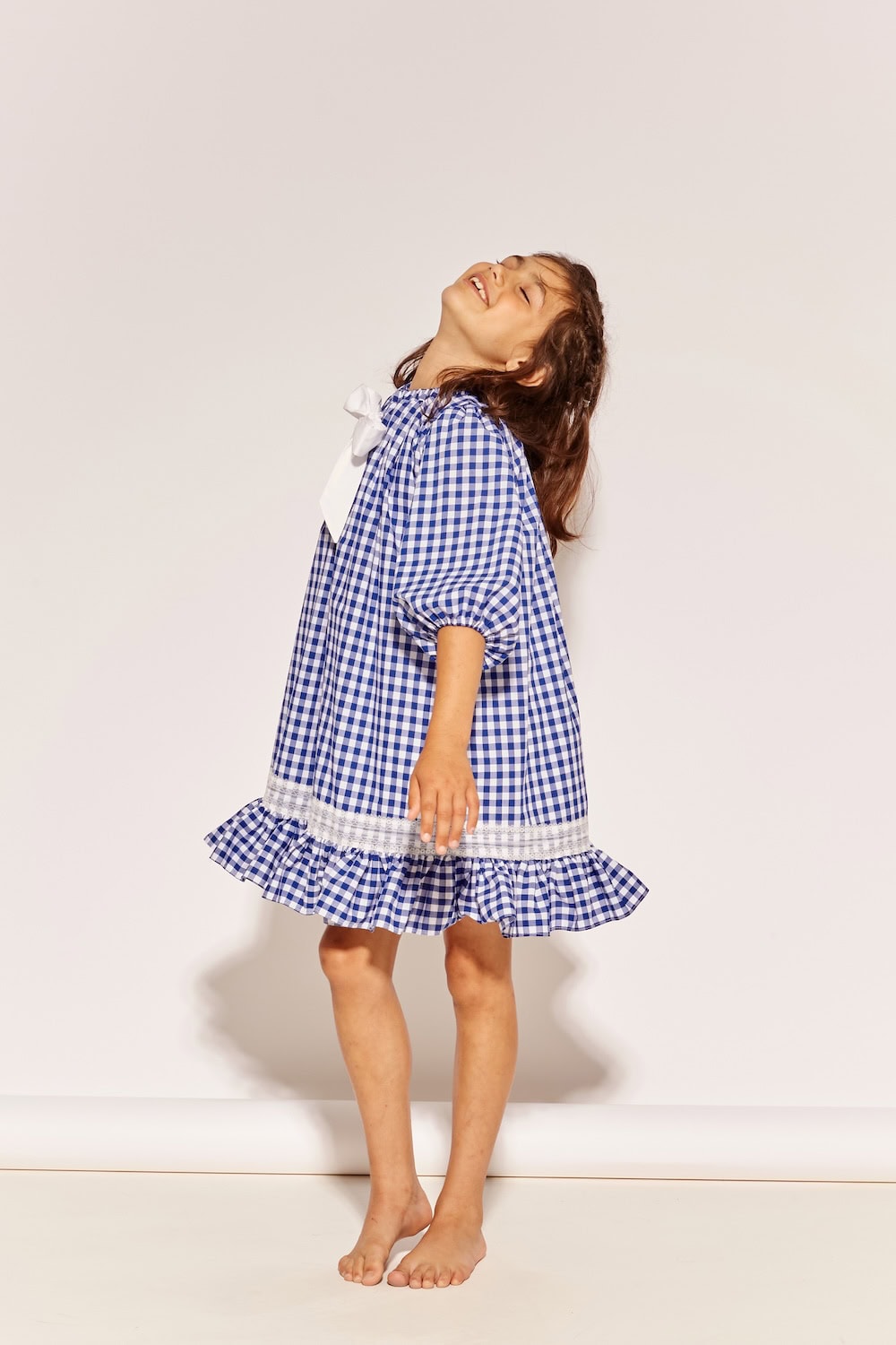 The Middle Daughter SS26 blue and white gingham dress with oversized white bow and ruffle hem on young girl in studio. Scimparello Magazine.