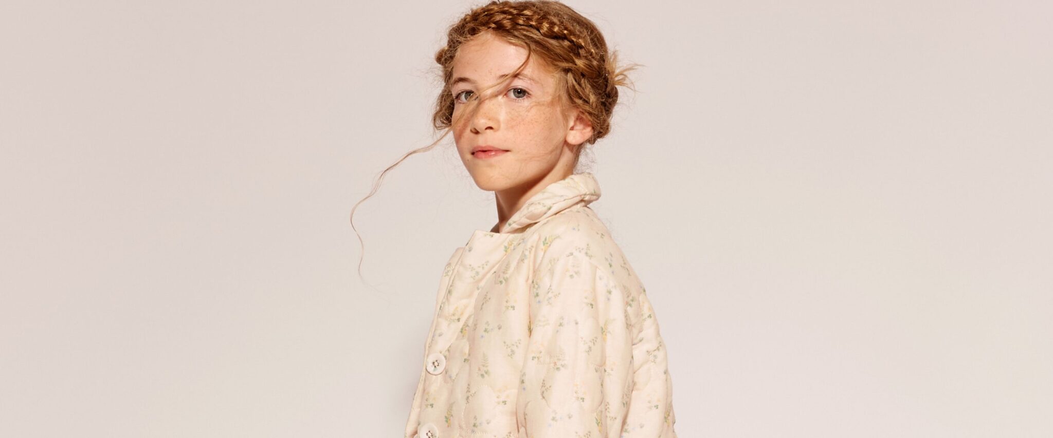 The Middle Daughter SS26 collection – portrait of a freckled girl with braided updo wearing a soft cream floral jacket with hood against a neutral background, delicate romantic styling. Scimparello Magazine.