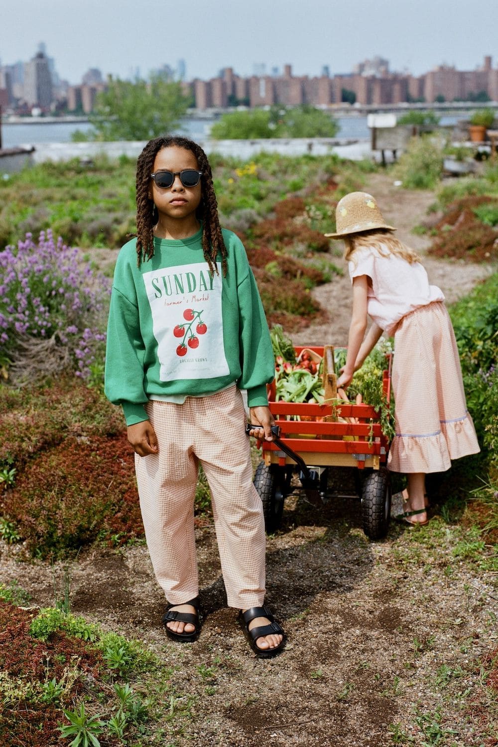 The Sunday Collective SS26 boy in green Sunday Farmers Market sweatshirt pulling red wagon in urban rooftop garden – Scimparello Magazine.
