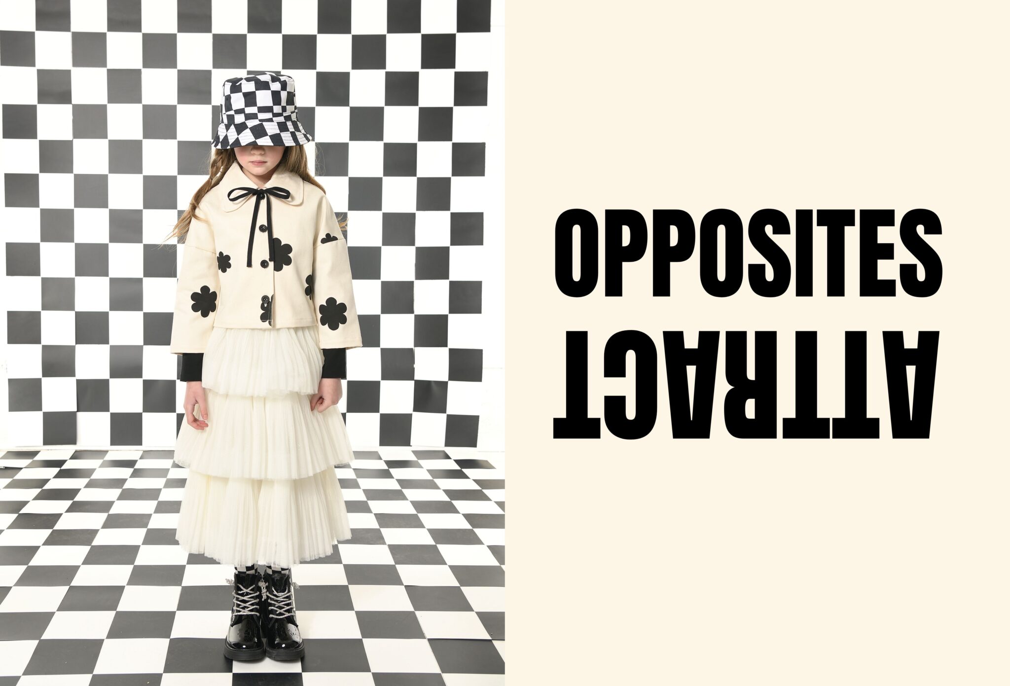 Girl wearing a cream layered tulle dress and matching jacket with black flower details, paired with a black-and-white checkered bucket hat and patent boots, standing in a graphic checkerboard set, kids fashion editorial SS26 Opposites Attract by David Butterman for Scimparello Magazine