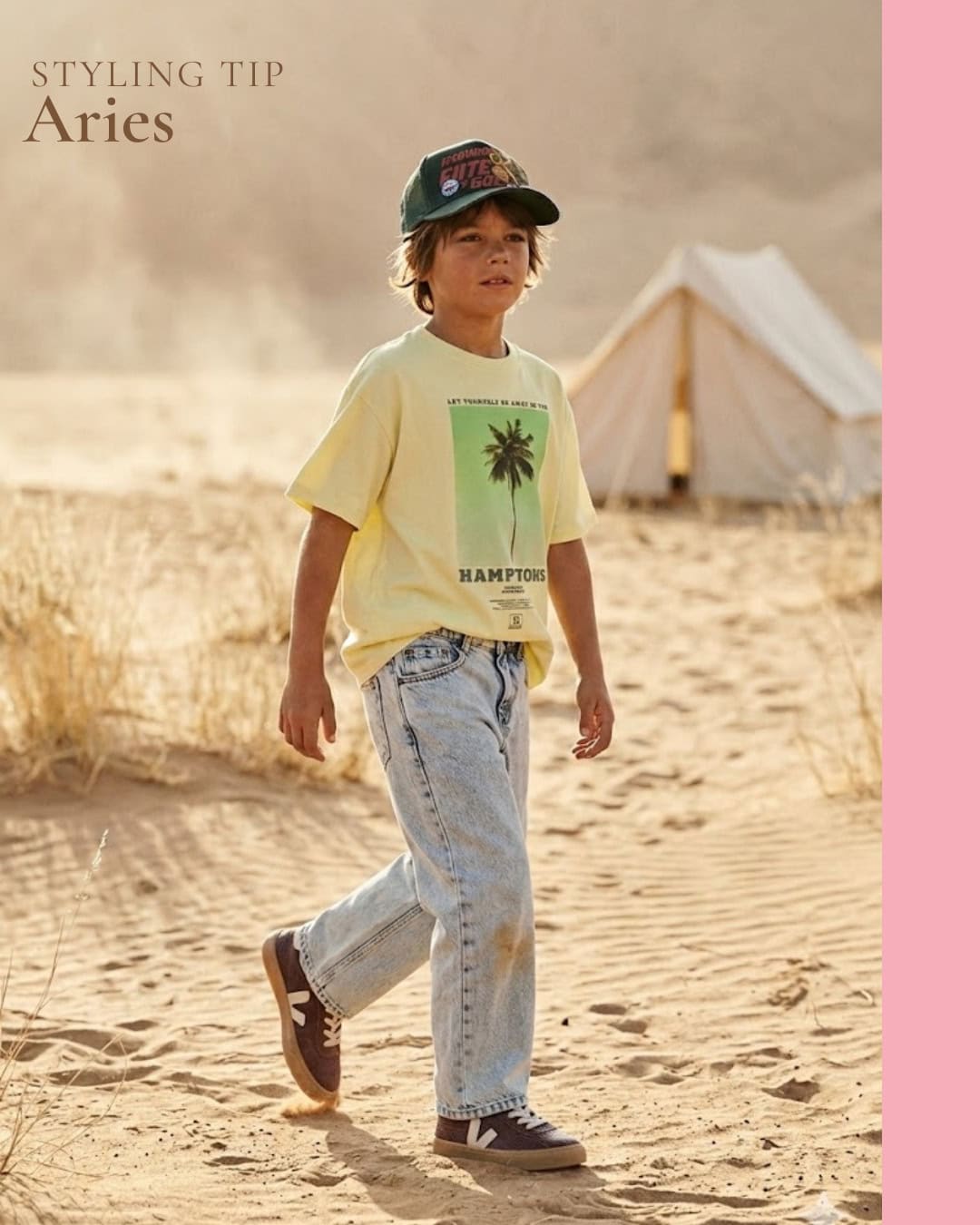 Scimparello Magazine Styling Tip Aries featuring a boy walking through sandy dunes at golden hour, wearing a pale yellow graphic t-shirt, light wash denim jeans, burgundy sneakers and a green cap, desert landscape with tent in the background, warm cinematic light