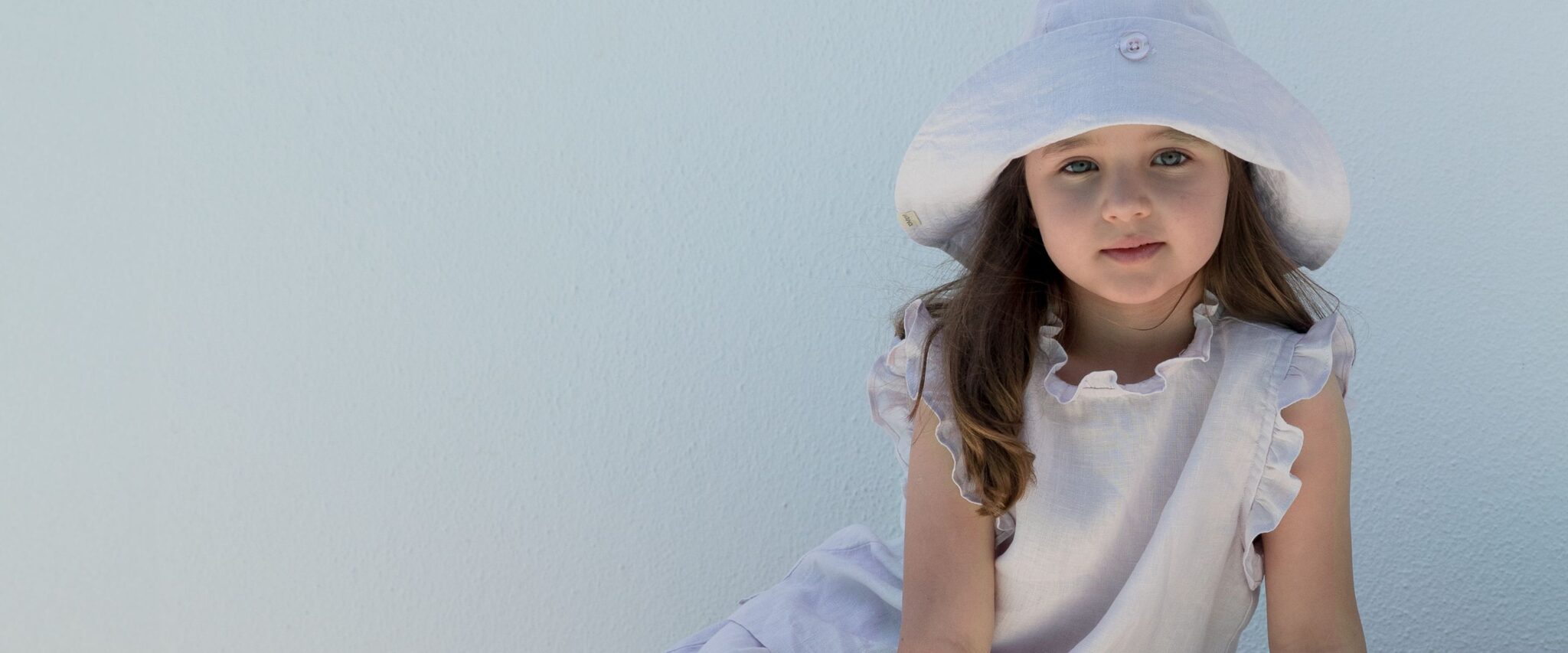 MarMar Copenhagen SS26 girl wearing a soft white summer dress with ruffled collar and wide white sun hat, sitting against a pale wall in natural light – kids fashion campaign image, Scimparello Magazine