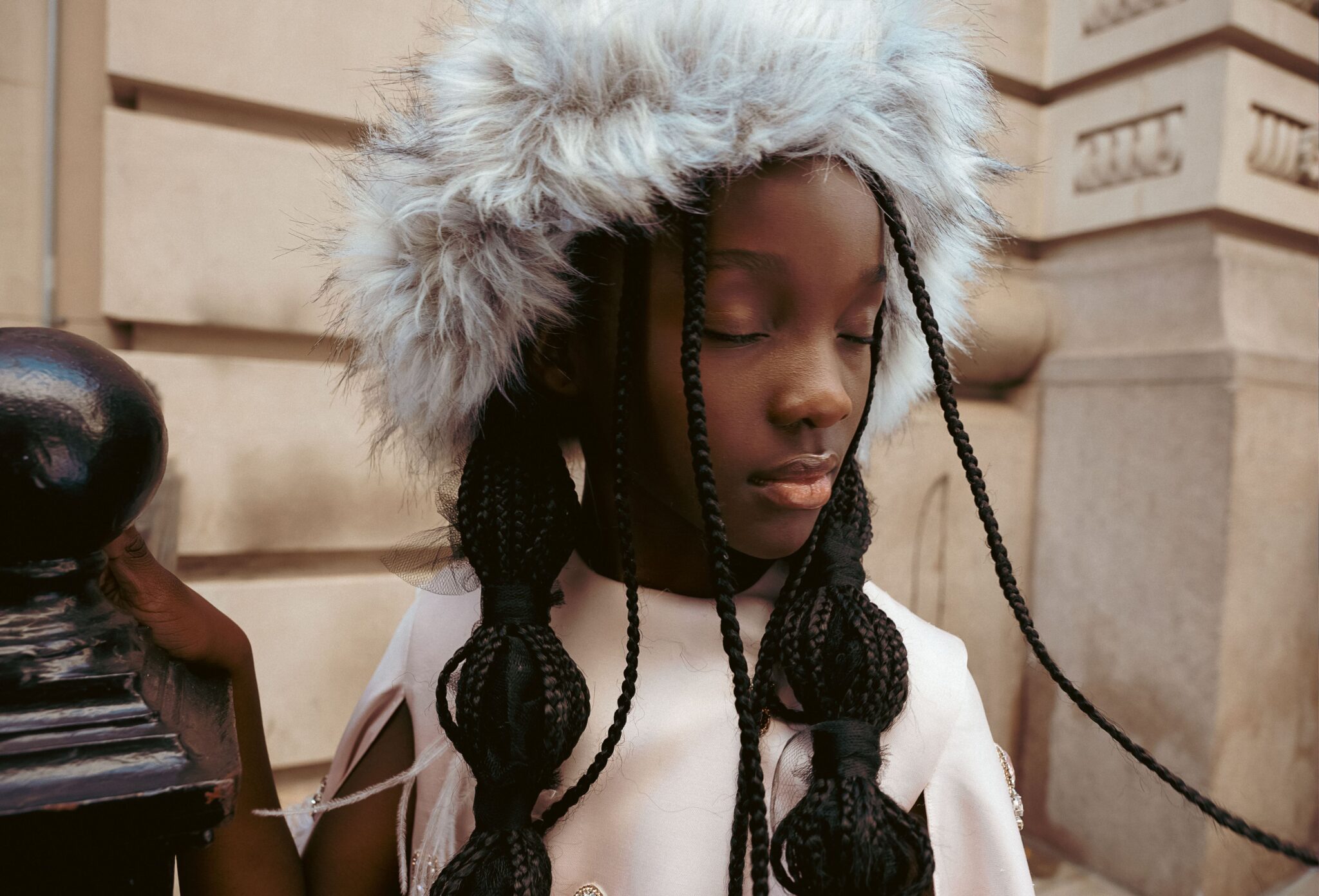 Kids fashion editorial The City Above Us for Scimparello Magazine. Portrait of a girl with long braids wearing a white embellished dress and soft grey fur hat, standing beside an iron railing in a New York street setting. Photo by Ilaria and Silvia Cianciaruso.