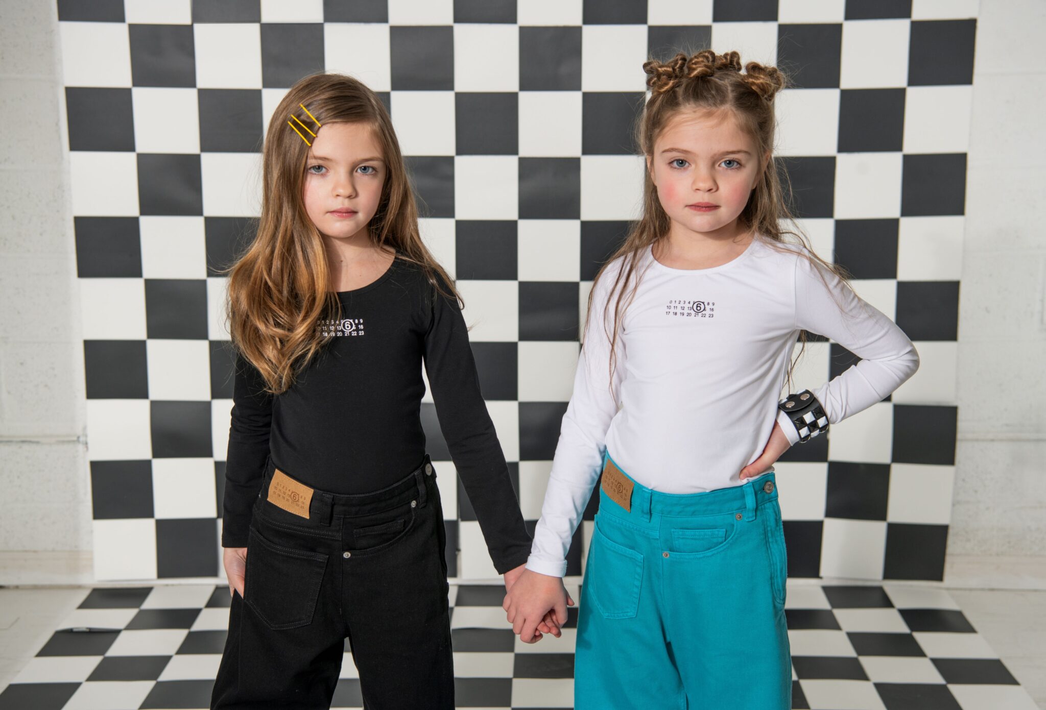 Two girls holding hands in a graphic black-and-white checkerboard set, one dressed in a black long-sleeve top with matching black pants and yellow hair clips, the other in a white long-sleeve top with teal pants and statement bracelet, kids fashion editorial SS26 Opposites Attract by David Butterman for Scimparello Magazine