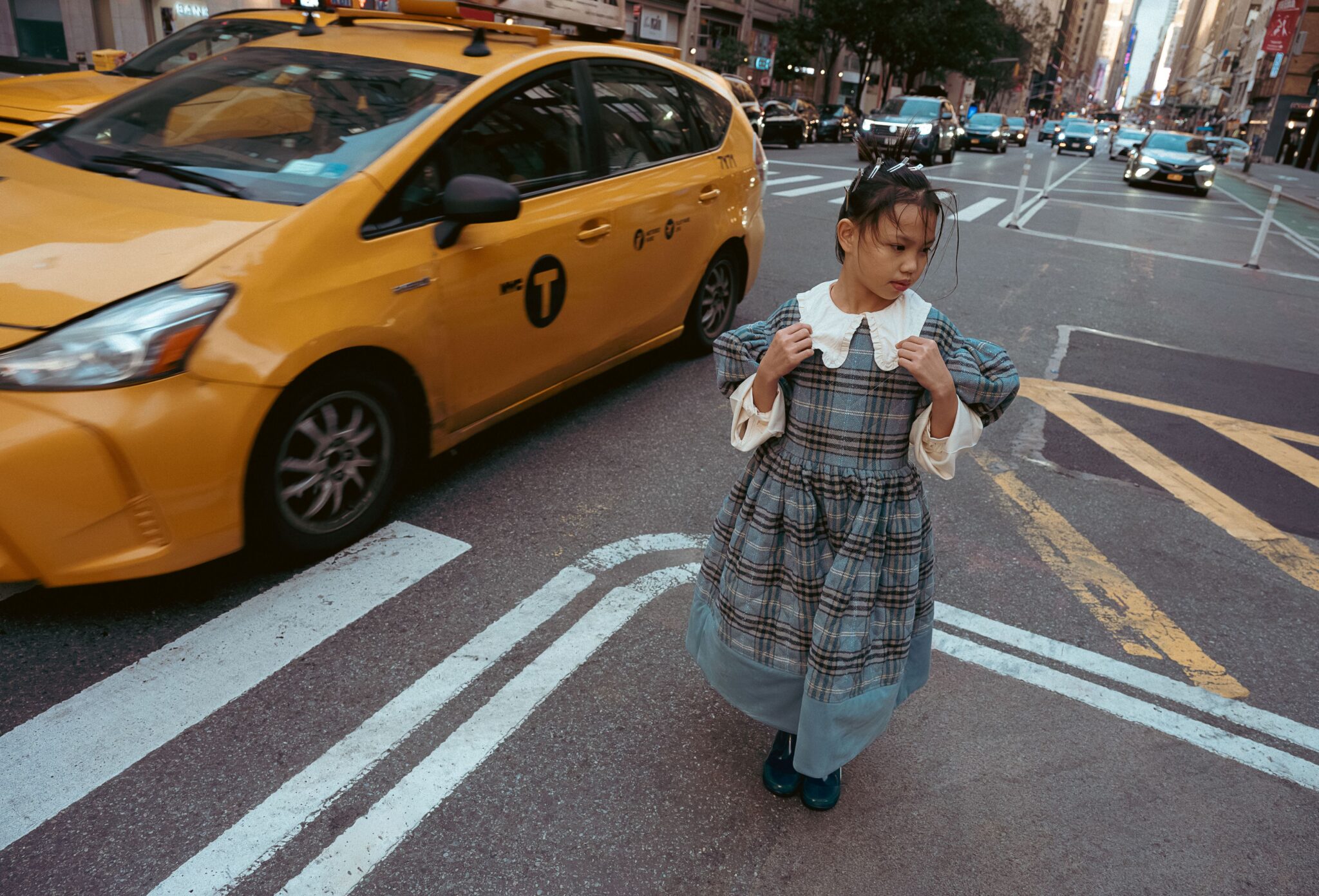 Kids fashion editorial The City Above Us for Scimparello Magazine. Girl wearing a plaid dress with a white collar walking across a New York street as a yellow taxi passes beside her, surrounded by city traffic and tall buildings. Photo by Ilaria and Silvia Cianciaruso.