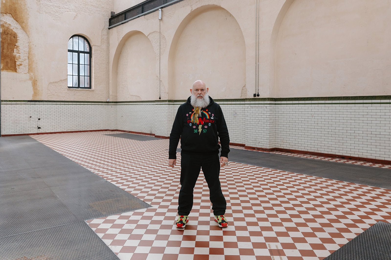 Portrait of designer Walter Van Beirendonck standing inside the C-mine industrial space, wearing a graphic hoodie and colorful sneakers on a checkered floor.