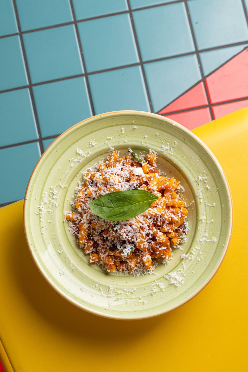 Fresh handmade pasta dish served in colorful plates at Fresca Pasta Club in Milan, featuring rich sauc topped with grated cheese and basil on a yellow table.