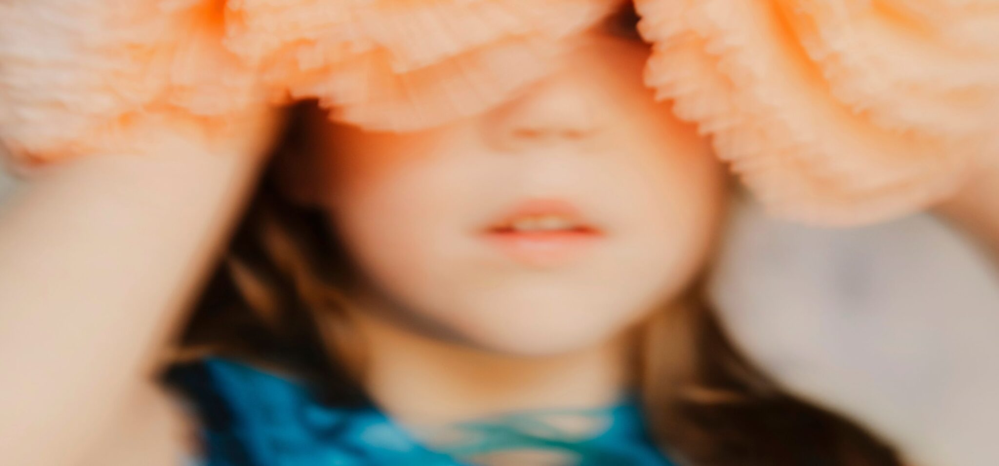 Soft-focus double spread from the kids fashion editorial Softly the Light Enters featuring a young girl in a flowing turquoise dress holding oversized peach tulle headpieces above her head, photographed by Hannah Lipowsky for Scimparello Magazine.