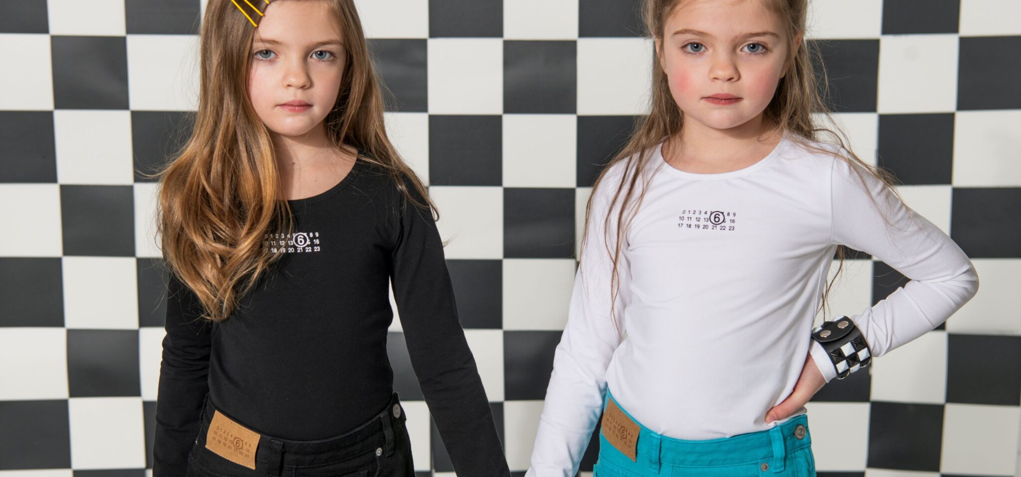 Two girls holding hands in a graphic black-and-white checkerboard set, one dressed in a black long-sleeve top with matching black pants and yellow hair clips, the other in a white long-sleeve top with teal pants and statement bracelet, kids fashion editorial SS26 Opposites Attract by David Butterman for Scimparello Magazine