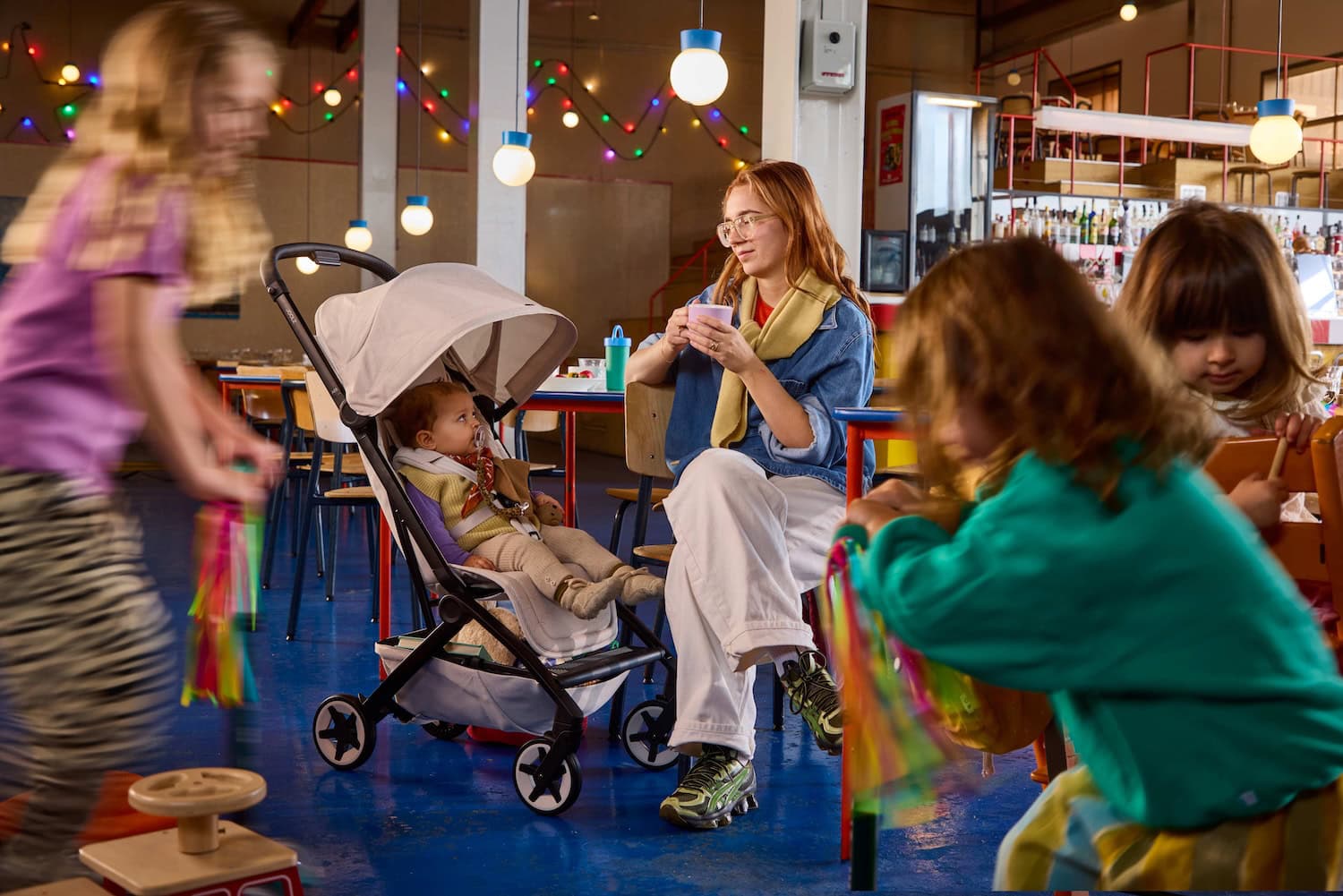 Parent sitting in a colorful café with a baby in the Joolz Aer2 Calming Beige stroller, surrounded by playful children, showcasing calm and comfort in a lively environment.