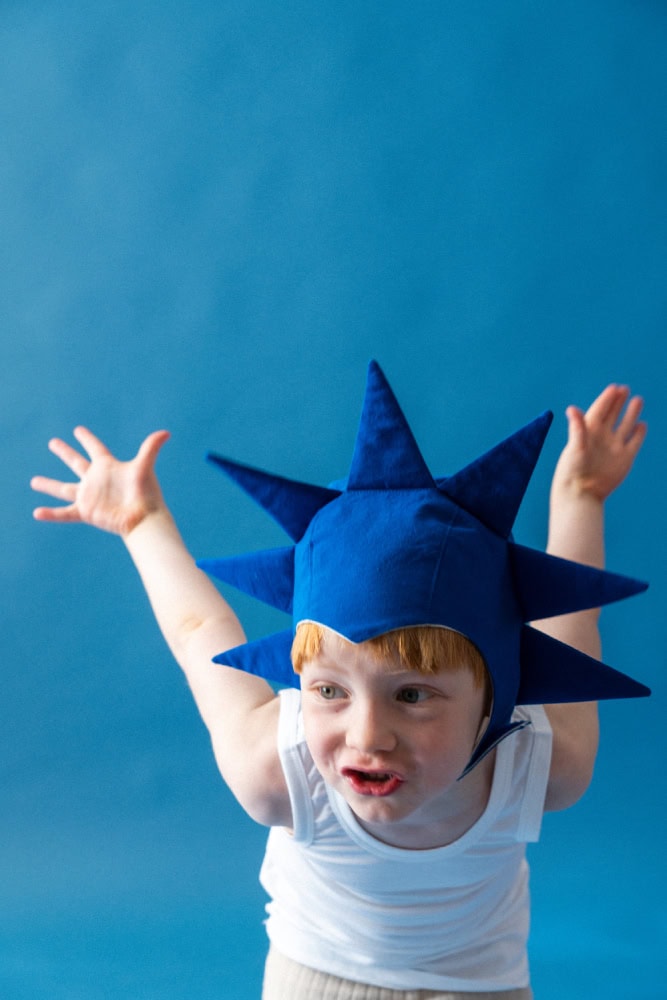 Child wearing a blue geometric star-shaped bonnet, raising arms playfully against a minimalist blue background