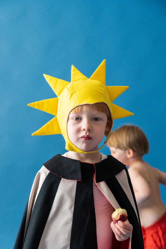 Child in a yellow sun-shaped bonnet and striped cape holding a bitten apple, with another child blurred in the background
