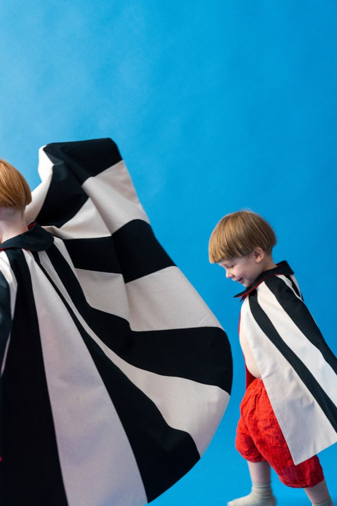 Two children wearing graphic capes and paper crowns, playing together in bold red, blue, black and white costumes
