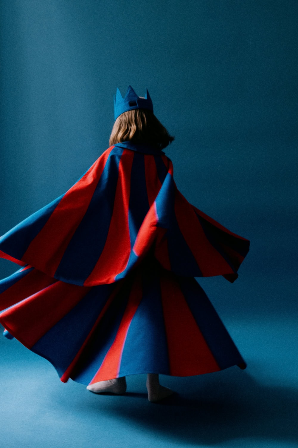 Child wearing a red and blue striped cape and crown, spinning dramatically against a deep blue background
