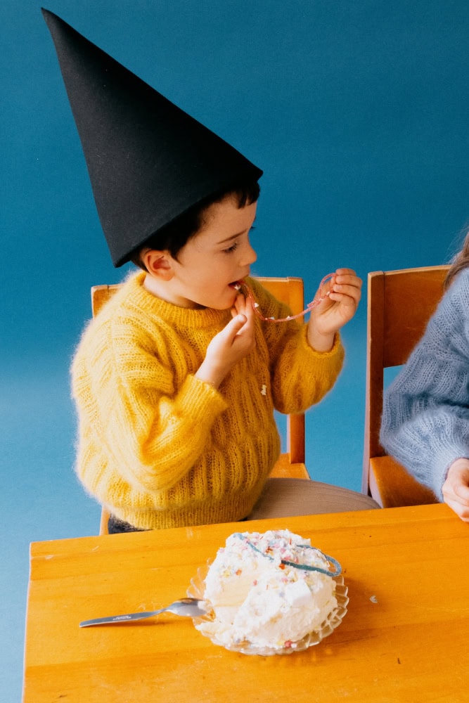 Child in a black pointed hat and yellow knit sweater eating cake at a wooden table, playful modern costume styling